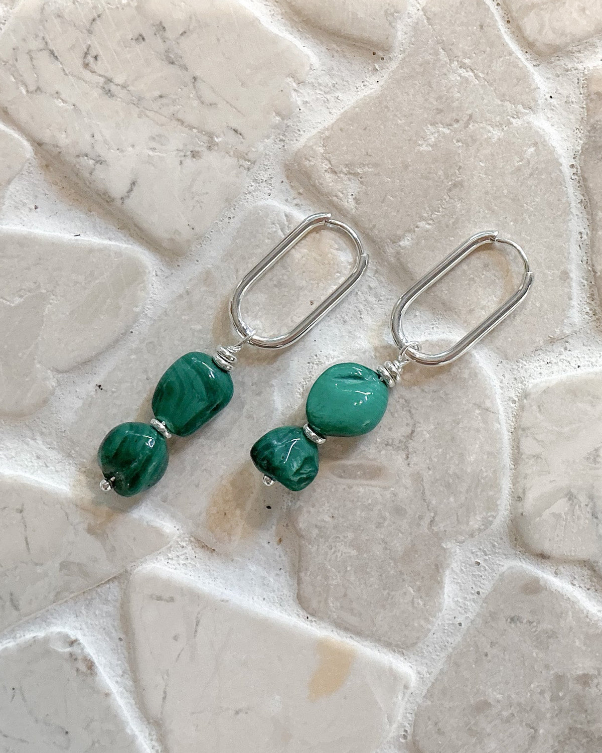 A pair of silver dangle earrings with green malachite beads, displayed on a light beige stone tile background.