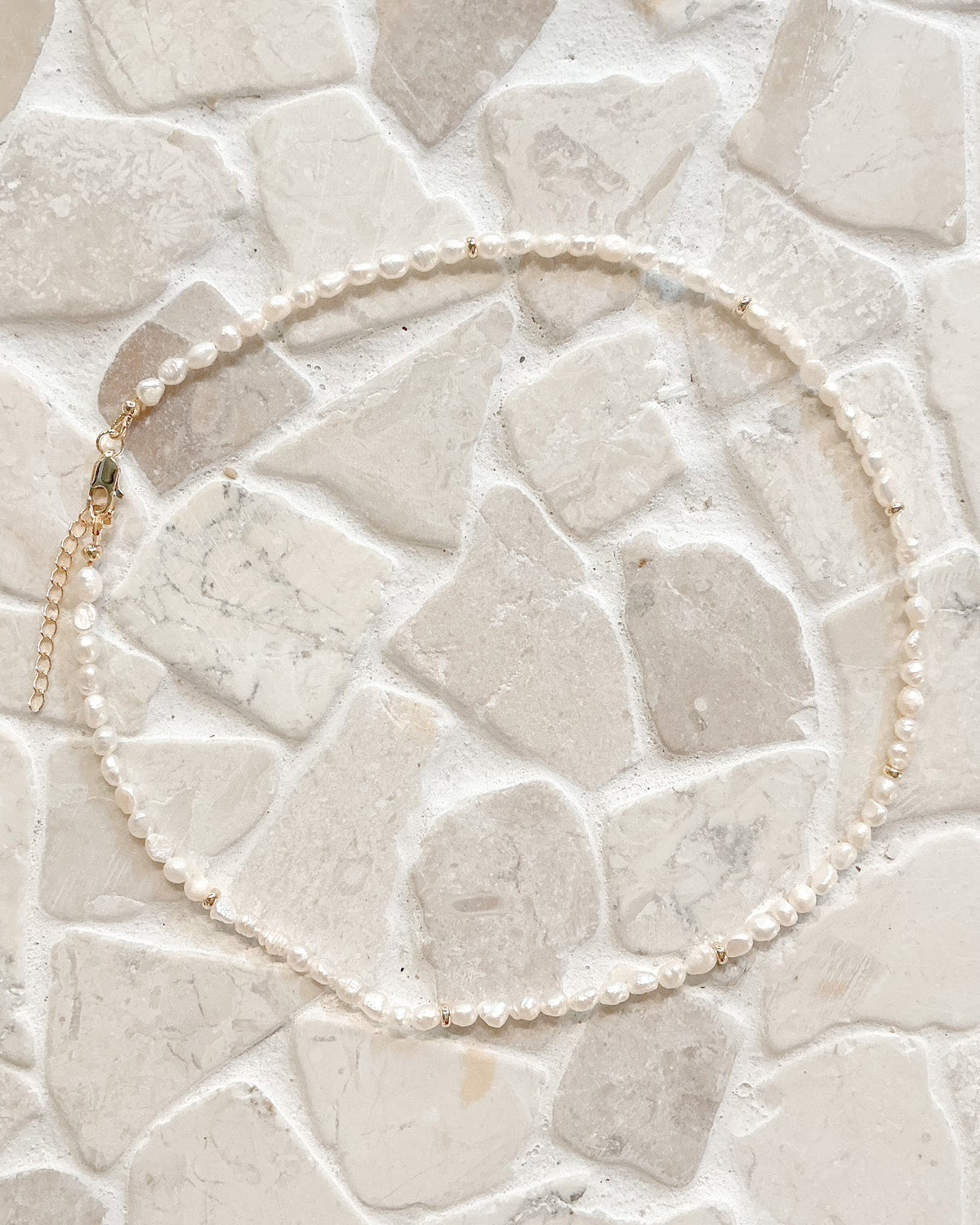 A delicate pearl necklace with a gold clasp, arranged in a circle on a light beige stone tile background.