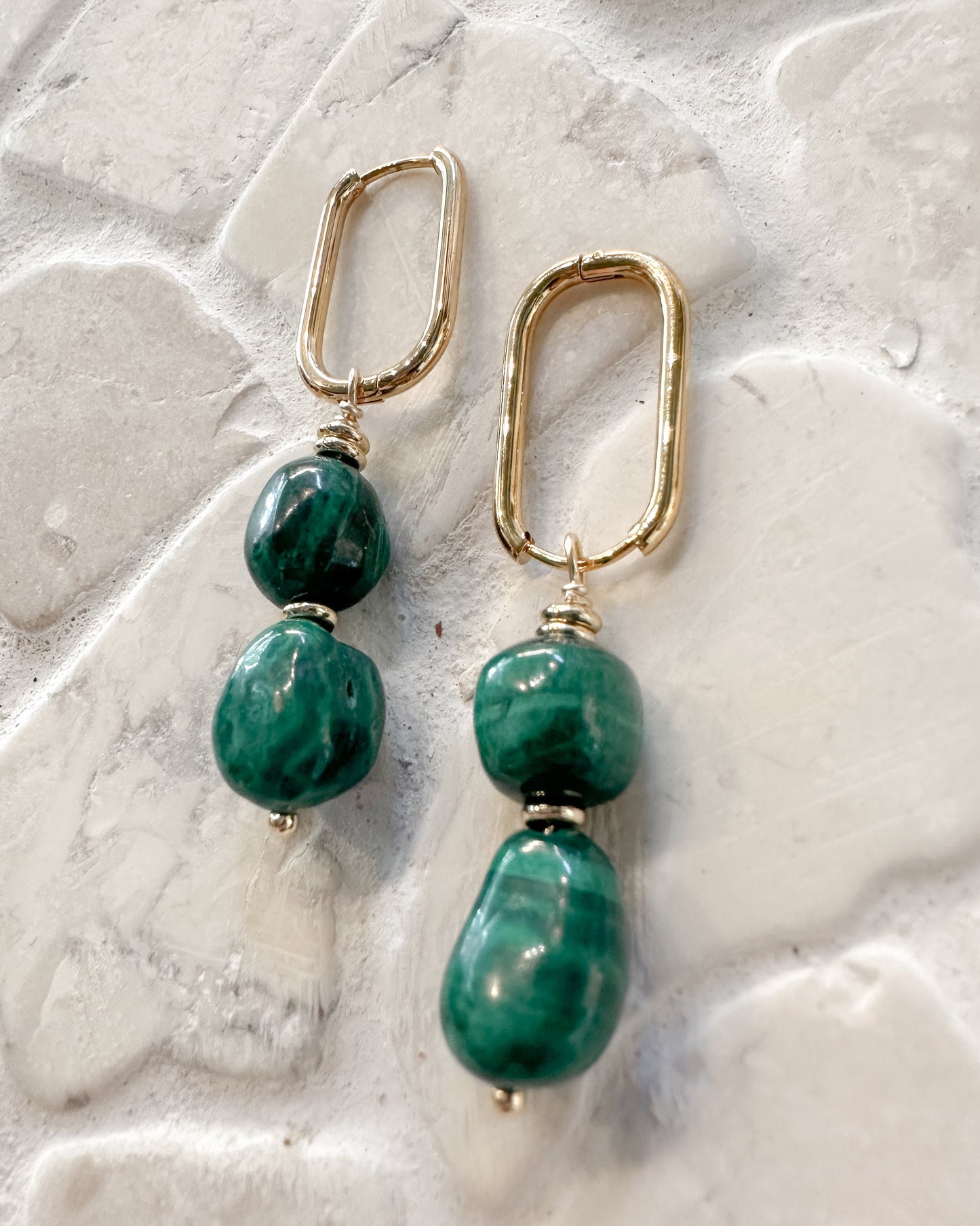 A pair of gold drop earrings with green marbled beads, displayed on a textured white stone surface.
