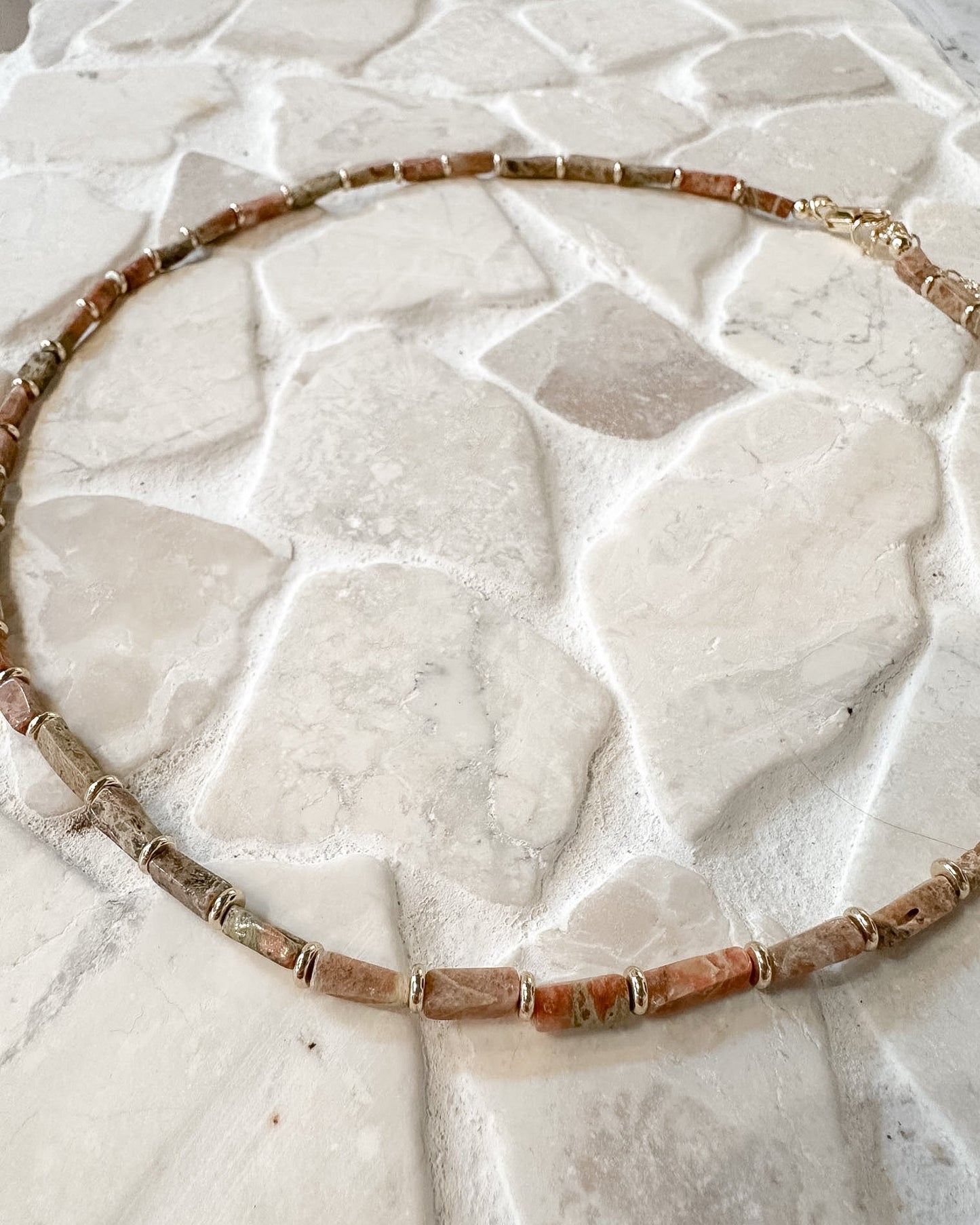 A beaded necklace made of terracotta and brown stone beads with small metallic spacer rings, laid flat on a light stone tile surface.