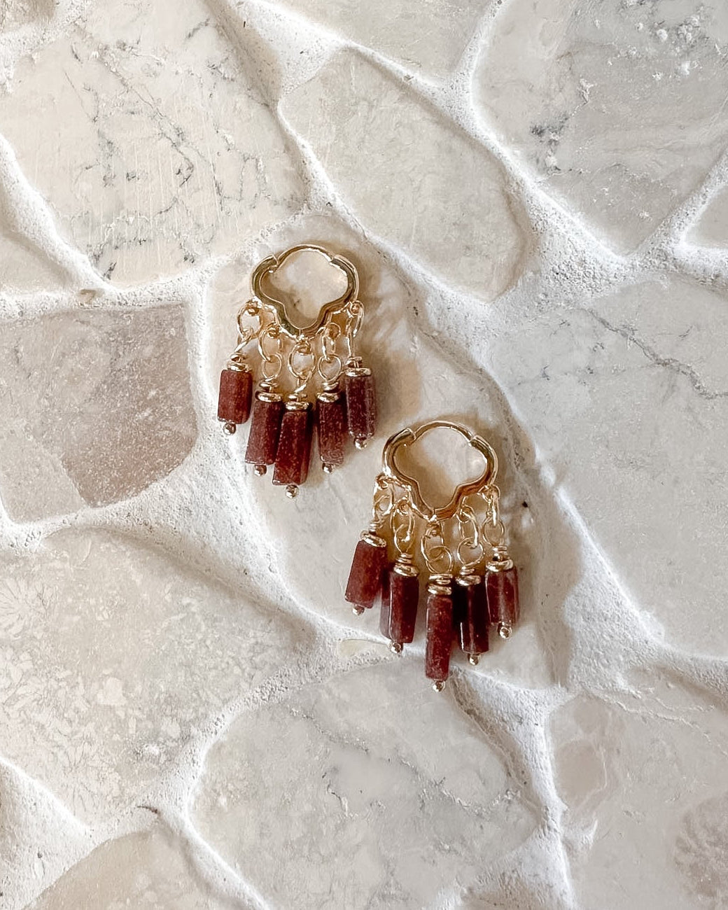 A pair of gold-toned dangle earrings with small clear beads and dangling dark red beads, placed on a textured white stone surface.