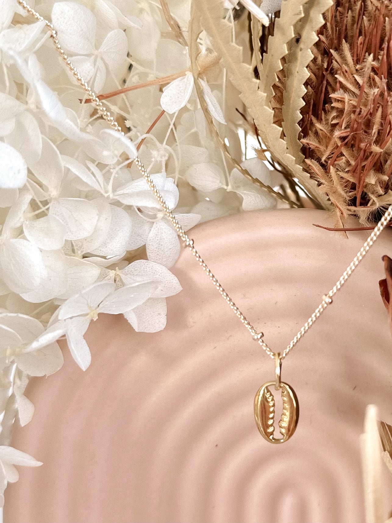Gold cowrie shell pendant on a gold chain draped over a pastel arched vase with dried flower