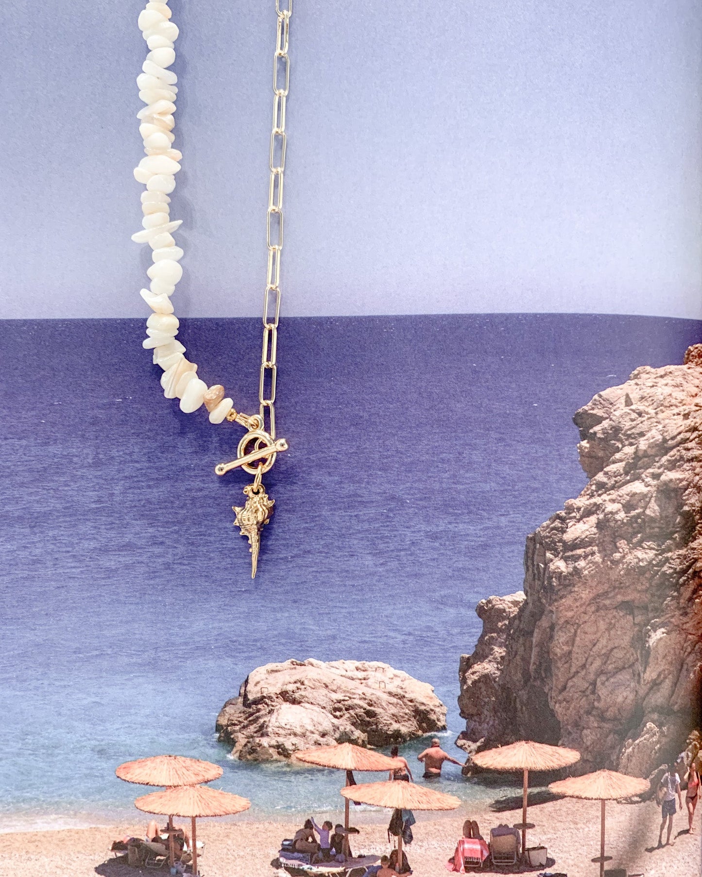A gold chain necklace with white shell-like beads and a gold anchor charm, shown against a split blue and lavender background with a beach scene printed along the left side.