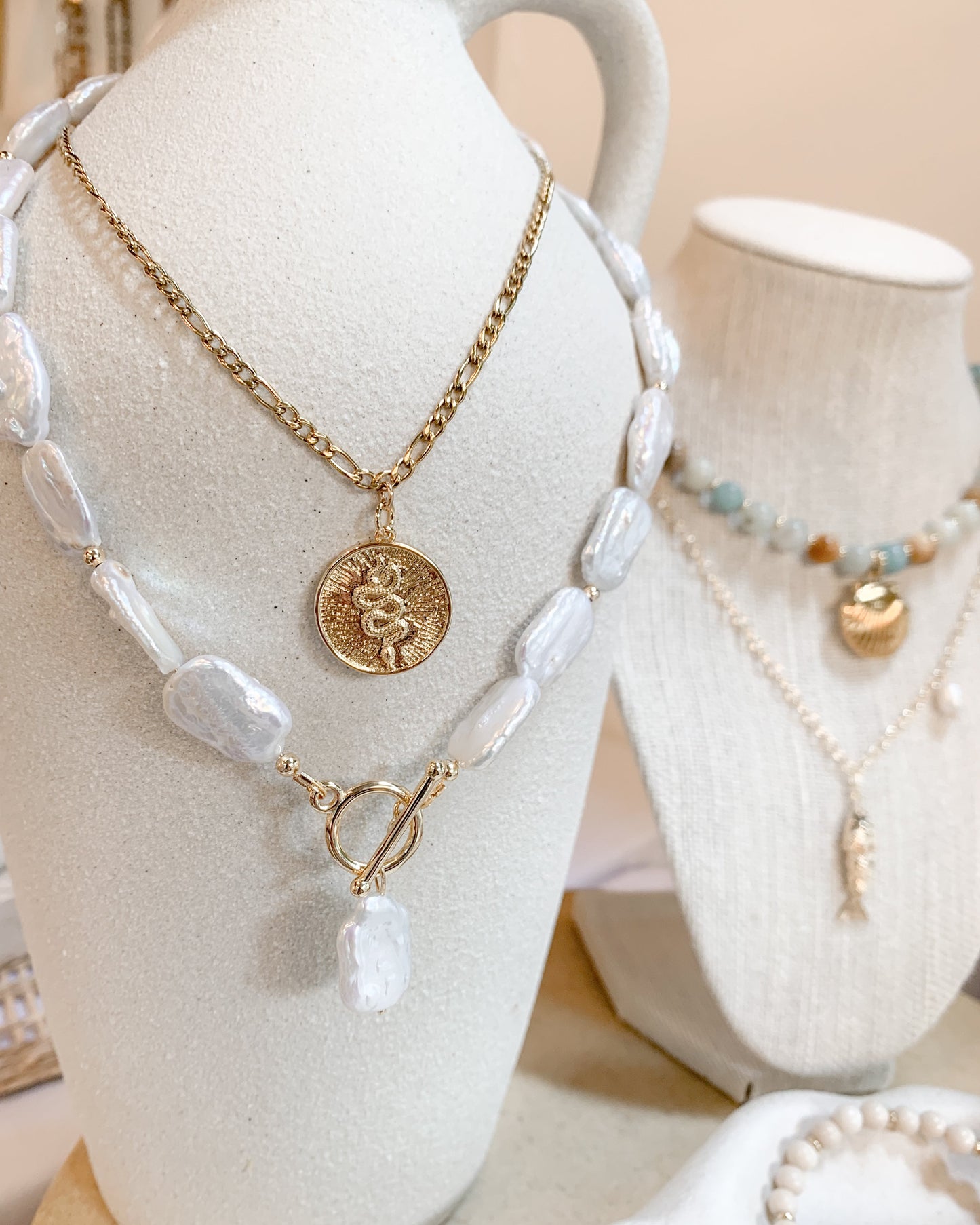 A gold chain necklace with freshwater baroque pearl beads and a round gold coin pendant displayed on a white ceramic pitcher. A second necklace with pale blue and white amazonite beads is partially visible on a second bust in the background.