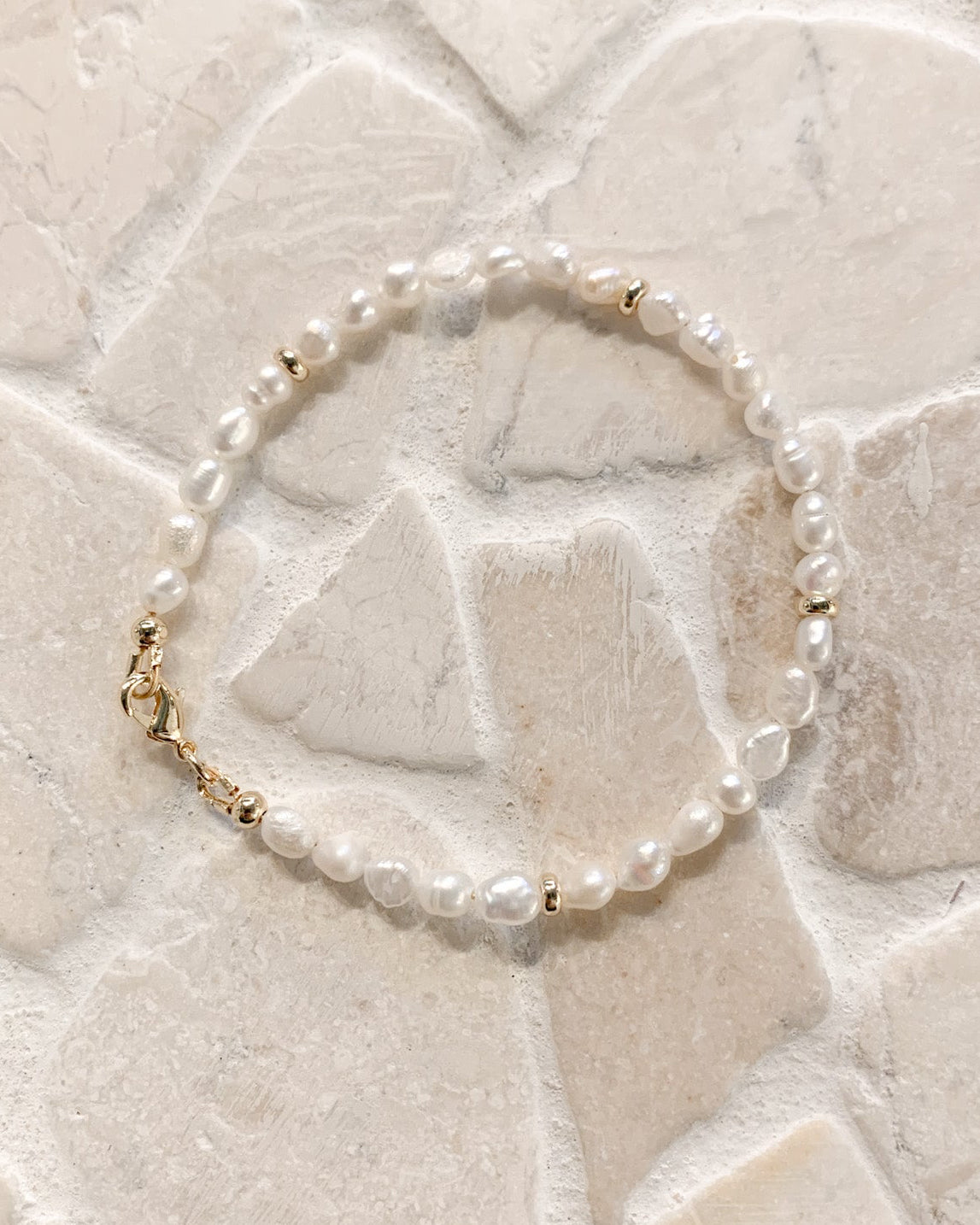 A delicate beaded bracelet with white freshwater pearls and small gold spacer beads, resting on a light beige stone tile background.
