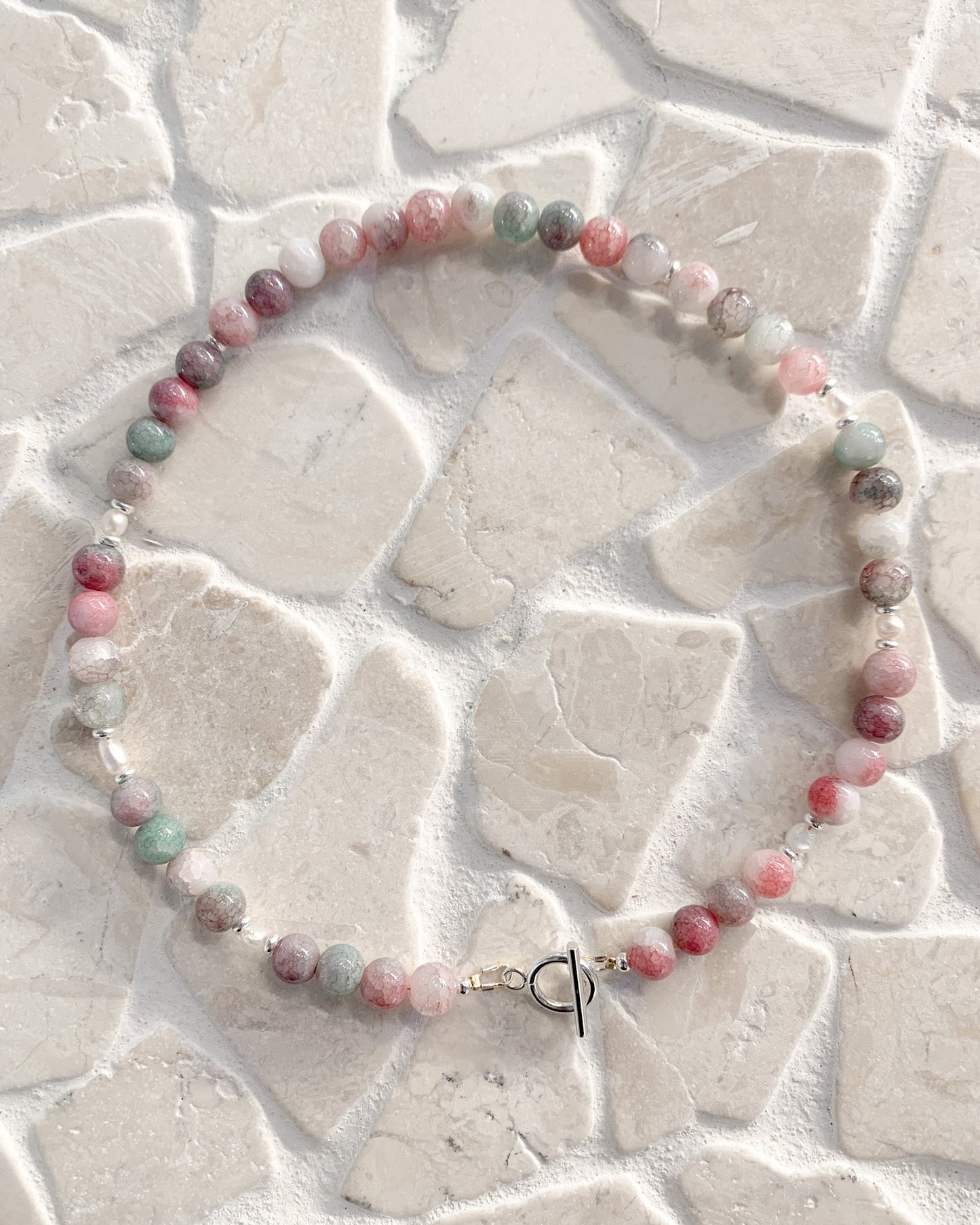 A beaded necklace made of round, polished gemstone beads in pink, green, gray, and white, arranged in a circle on a light beige stone tile background. The necklace features a small silver toggle clasp.