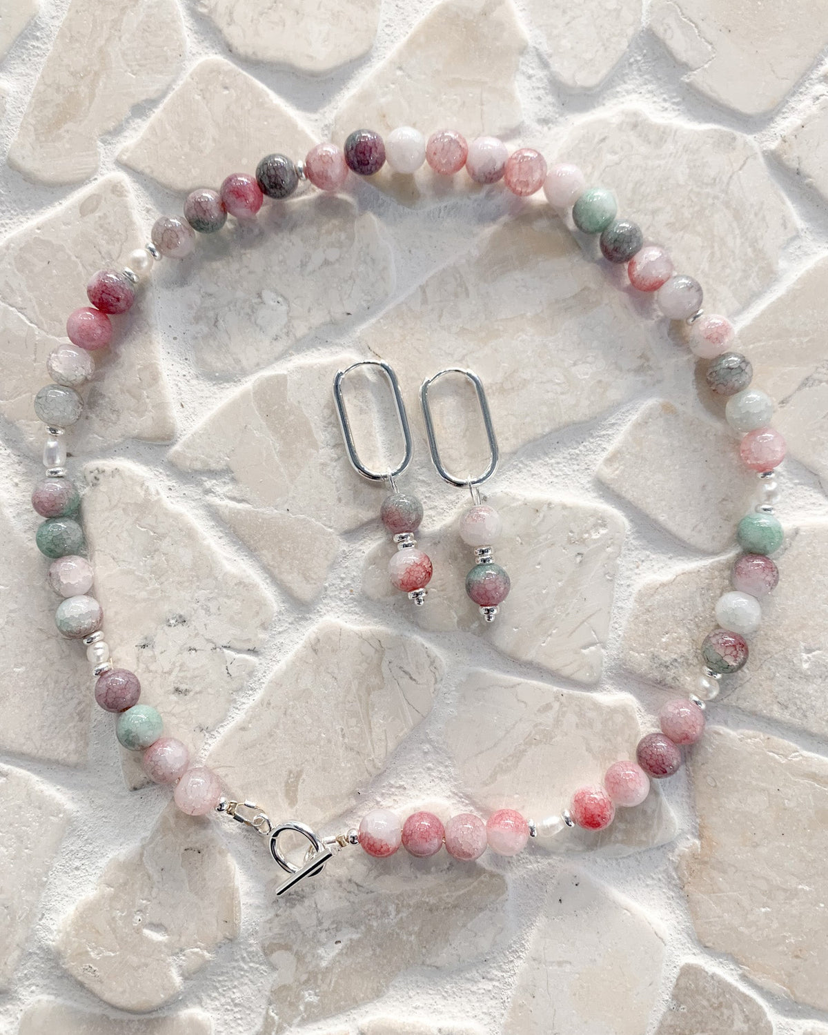 A beaded necklace and matching drop earrings laid flat on a light stone-textured surface. The jewelry features round beads in pink, white, mint green, and gray, with silver-toned metal findings including oval ear wires and a toggle clasp.