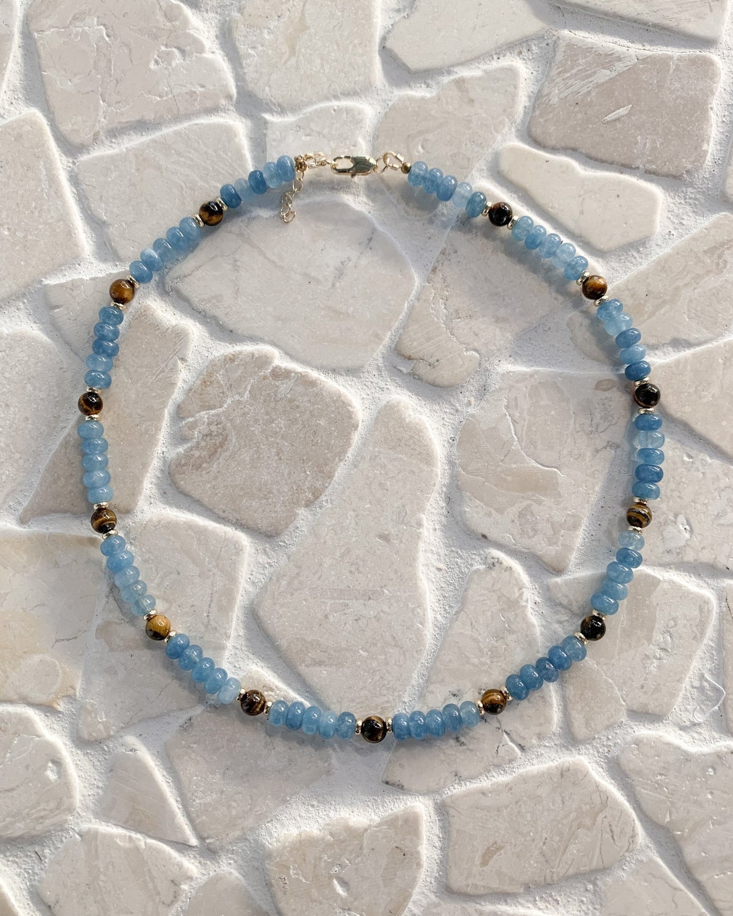 A beaded necklace with light blue faceted beads and small metallic gold spacer beads, arranged in a circle on a light beige stone tile background.