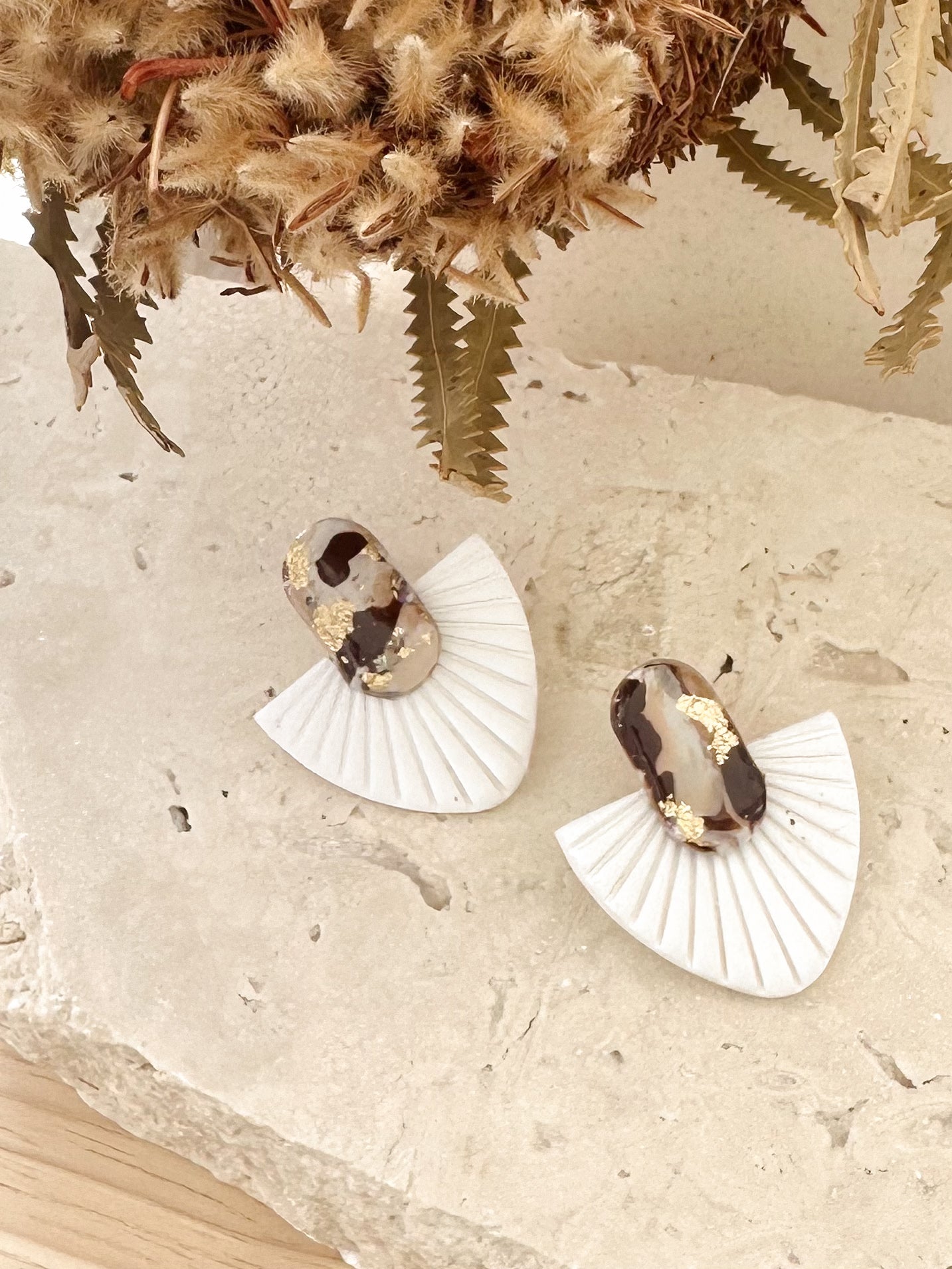 Pair of fan shaped sculptural earrings on a textured surface with dried plants in the background