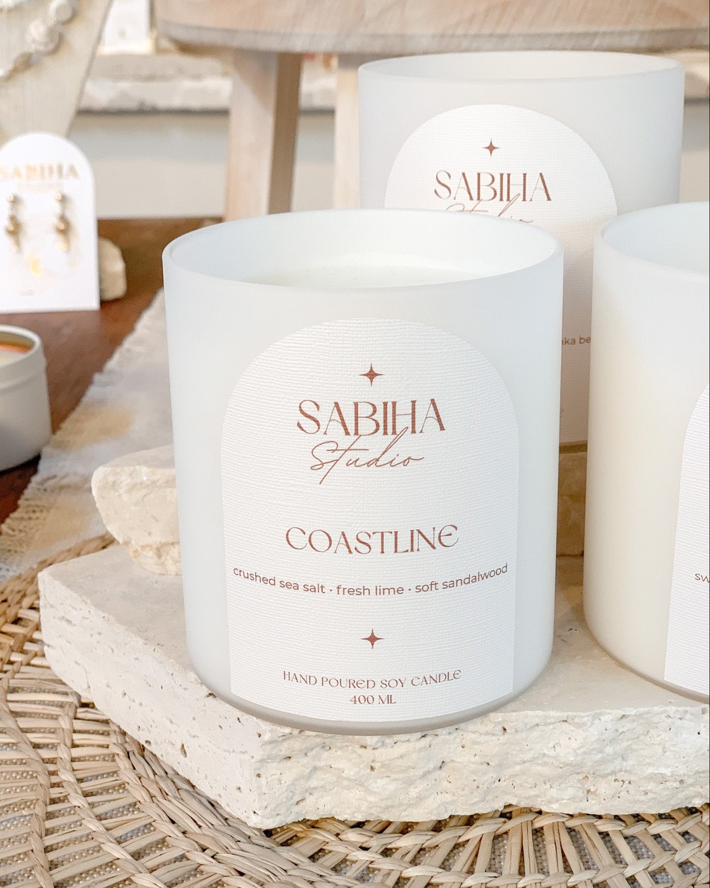A white soy candle jar labeled 'SABHIA Studios COASTLINE' sits on a woven tray with other matching candles and a small jewelry display in a softly styled indoor setting.