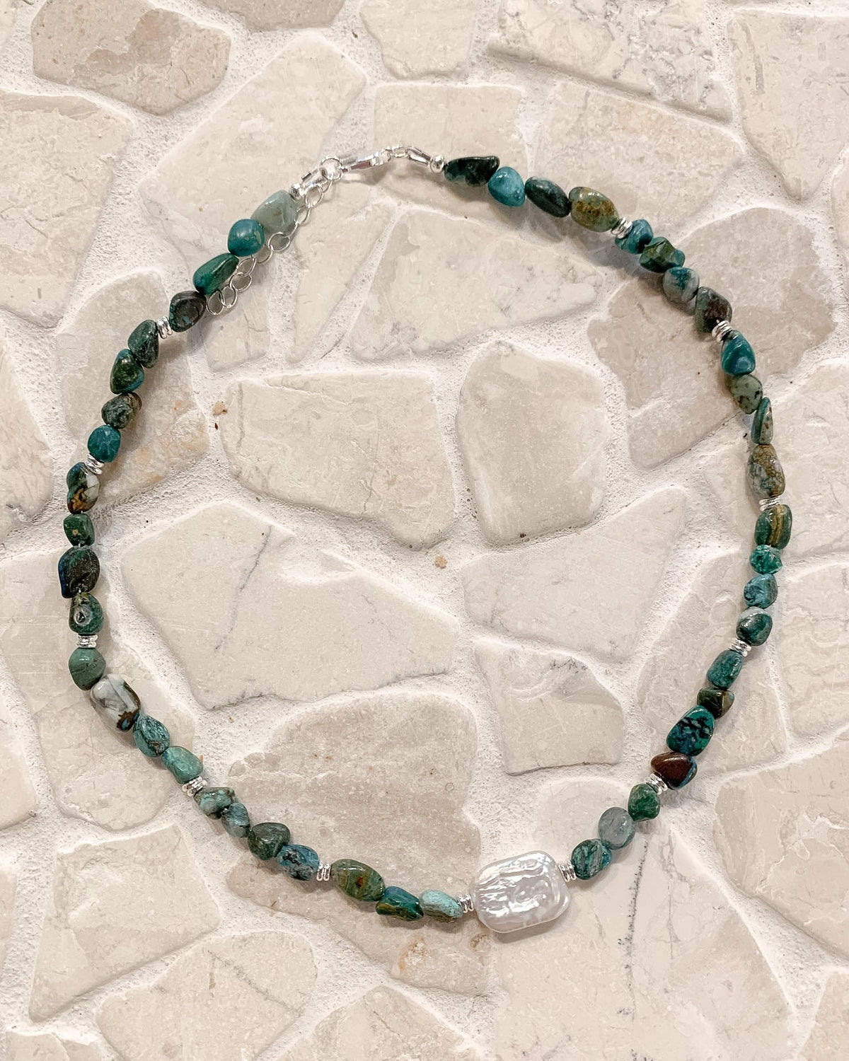 A beaded necklace made of polished green and blue stones with a clear faceted crystal pendant, displayed on a light beige stone tile background.