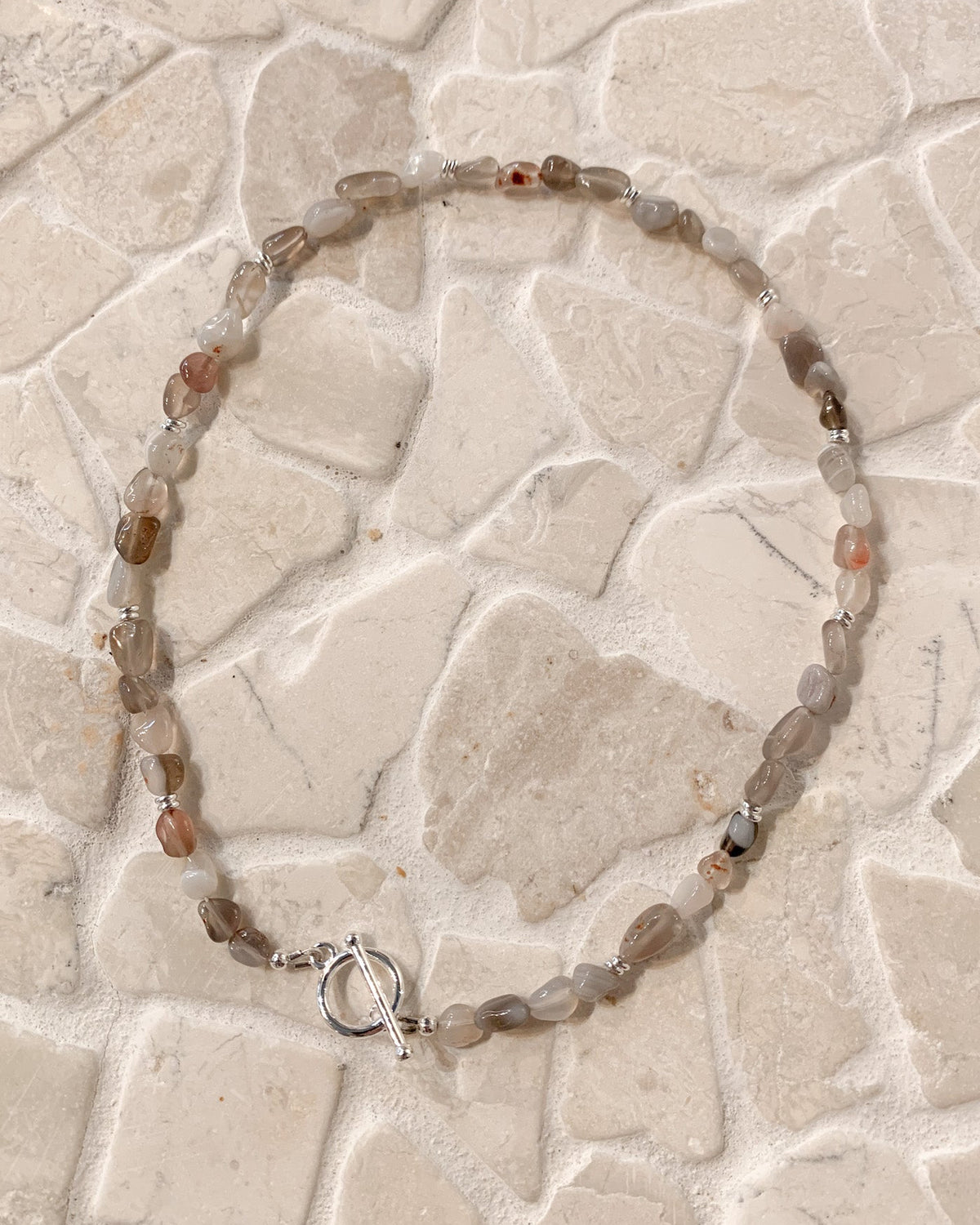 A beaded necklace made of irregular translucent gray and brown gemstone beads with a silver toggle clasp, laid flat on a light beige stone tile surface.