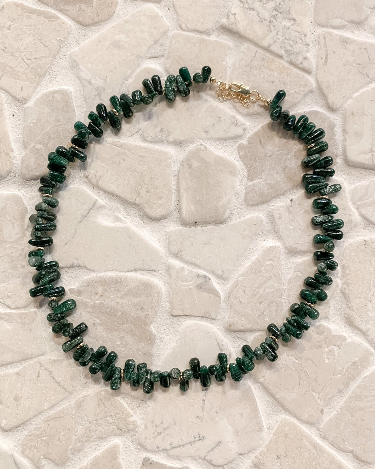 A beaded necklace made of small dark green stone chips with a gold clasp, displayed on a light beige stone tile surface.