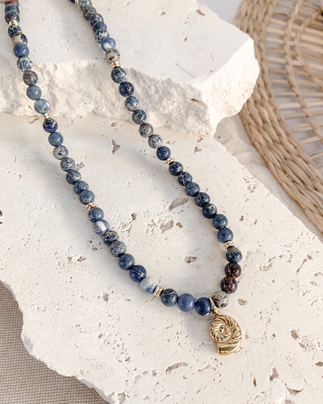 A beaded necklace with dark blue and grey marbled sodalite beads, gold spacer beads, and a small gold skull pendant, displayed on a white stone prop.