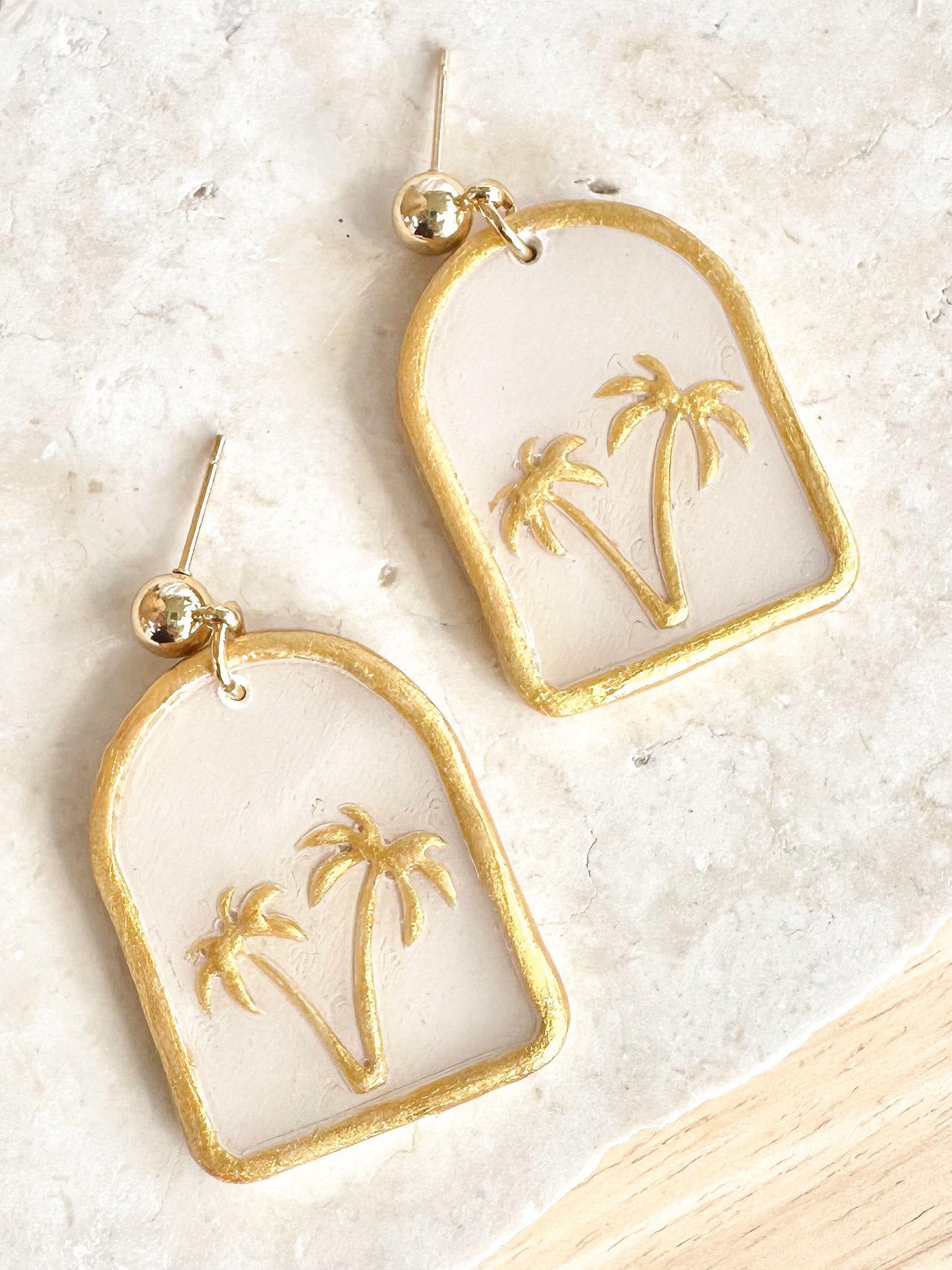 Gold and white earrings with palm tree design on a light background