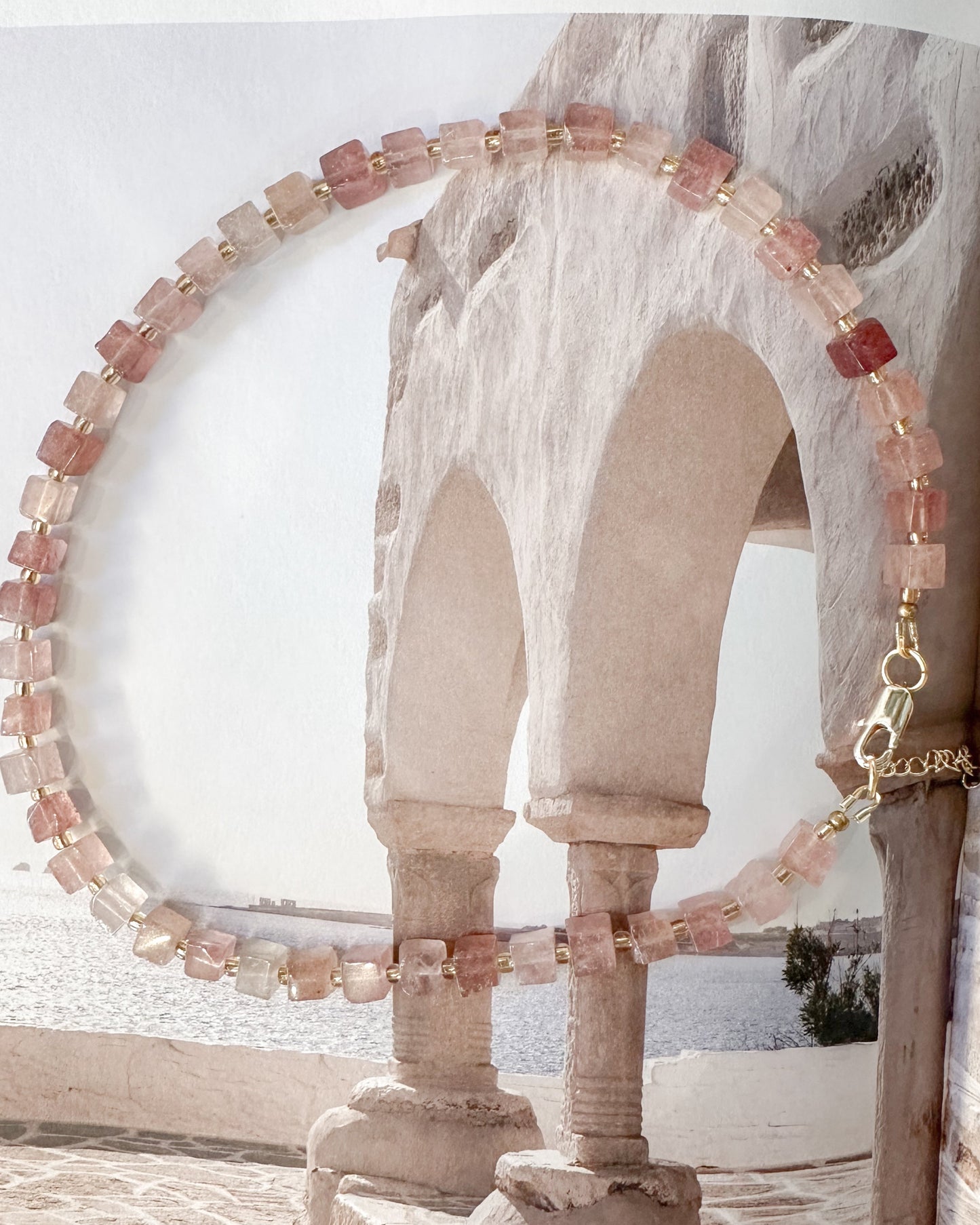 A beaded necklace with strawberry quartz square beads and a gold clasp, draped over an open book displaying a scenic photograph of stone arches.