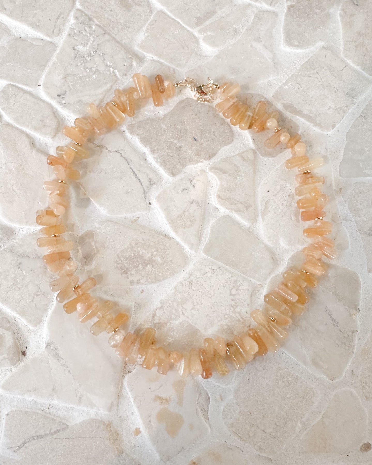 A beaded necklace made of small, irregular peach and orange stone chips, displayed on a textured white stone surface.
