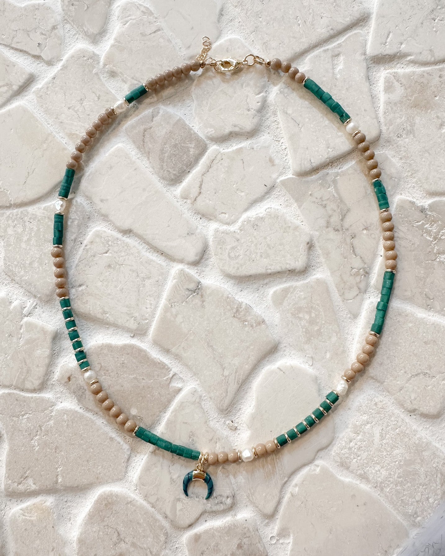 A beaded necklace with alternating strands of light beige beads and dark green beads, featuring a small gold crescent moon charm at the center, displayed on a textured white stone surface.