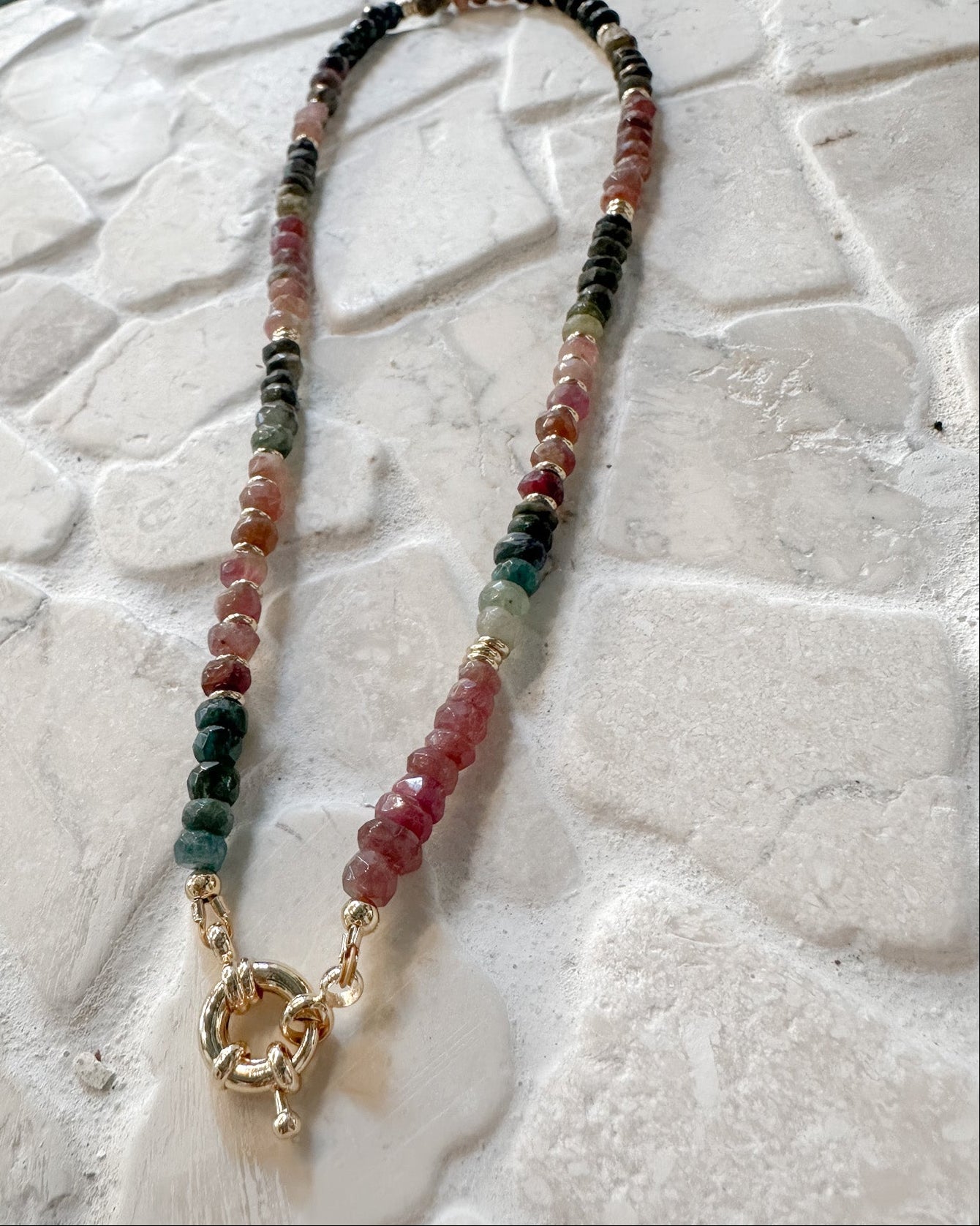 A beaded necklace made of small multicolored gemstone beads with a gold toggle clasp, displayed on a textured white stone surface.