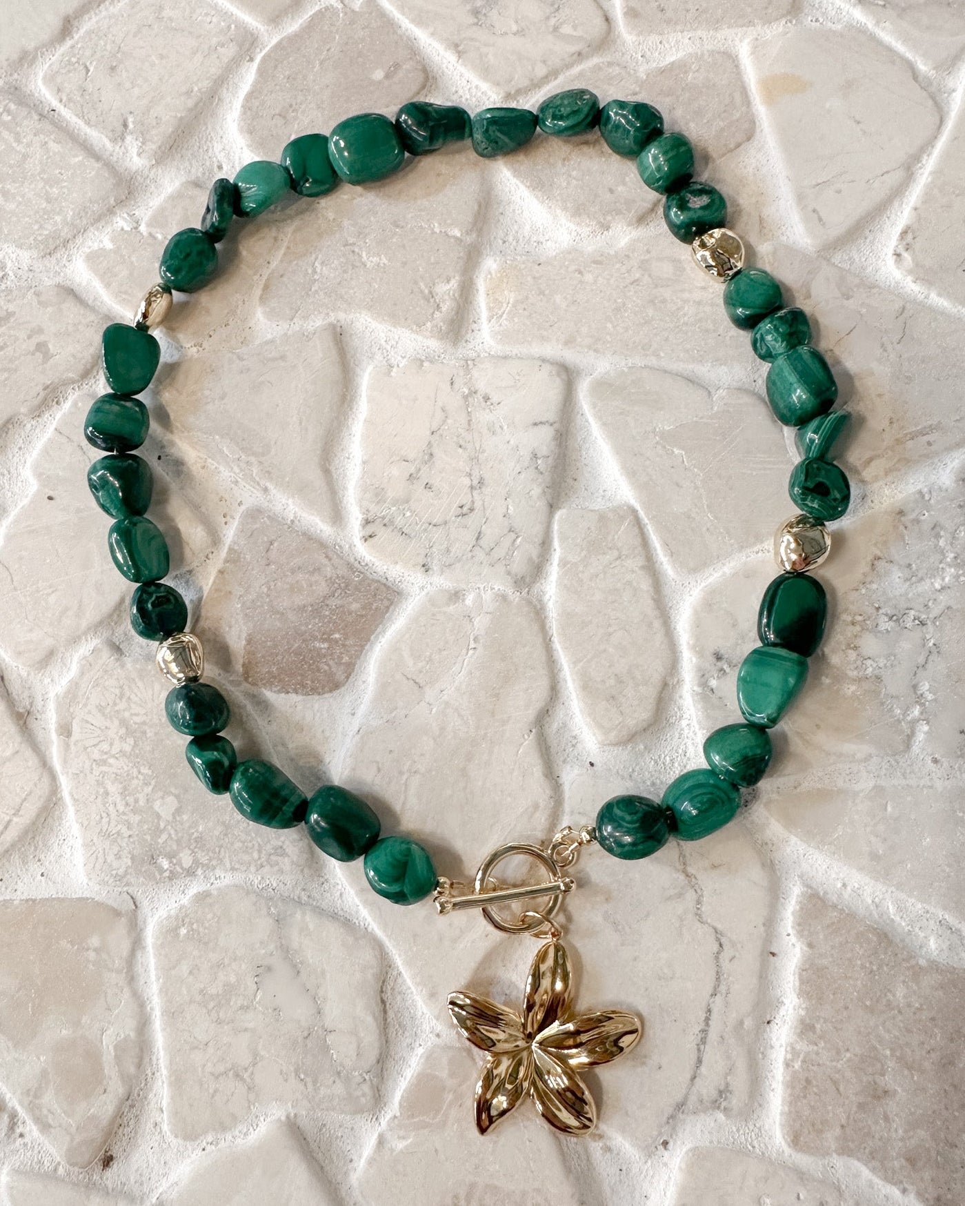 A beaded bracelet made of polished green malachite beads with a gold toggle clasp and a gold flower charm, displayed on a light beige stone tile background.