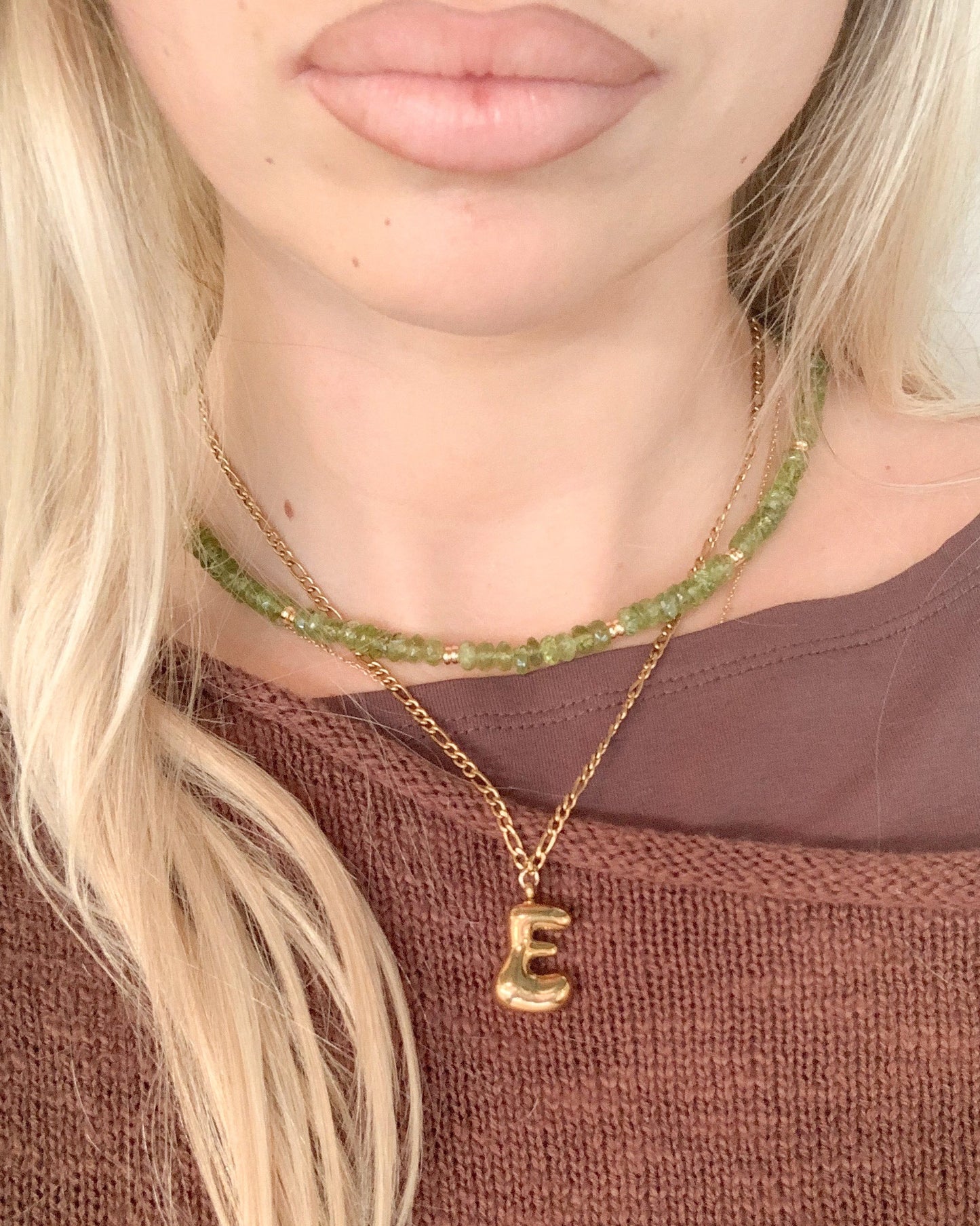 A close-up of a model wearing a layered necklace set. The set includes a gold chain with a gold bubble letter 'E' pendant and a green beaded peridot choker necklace.