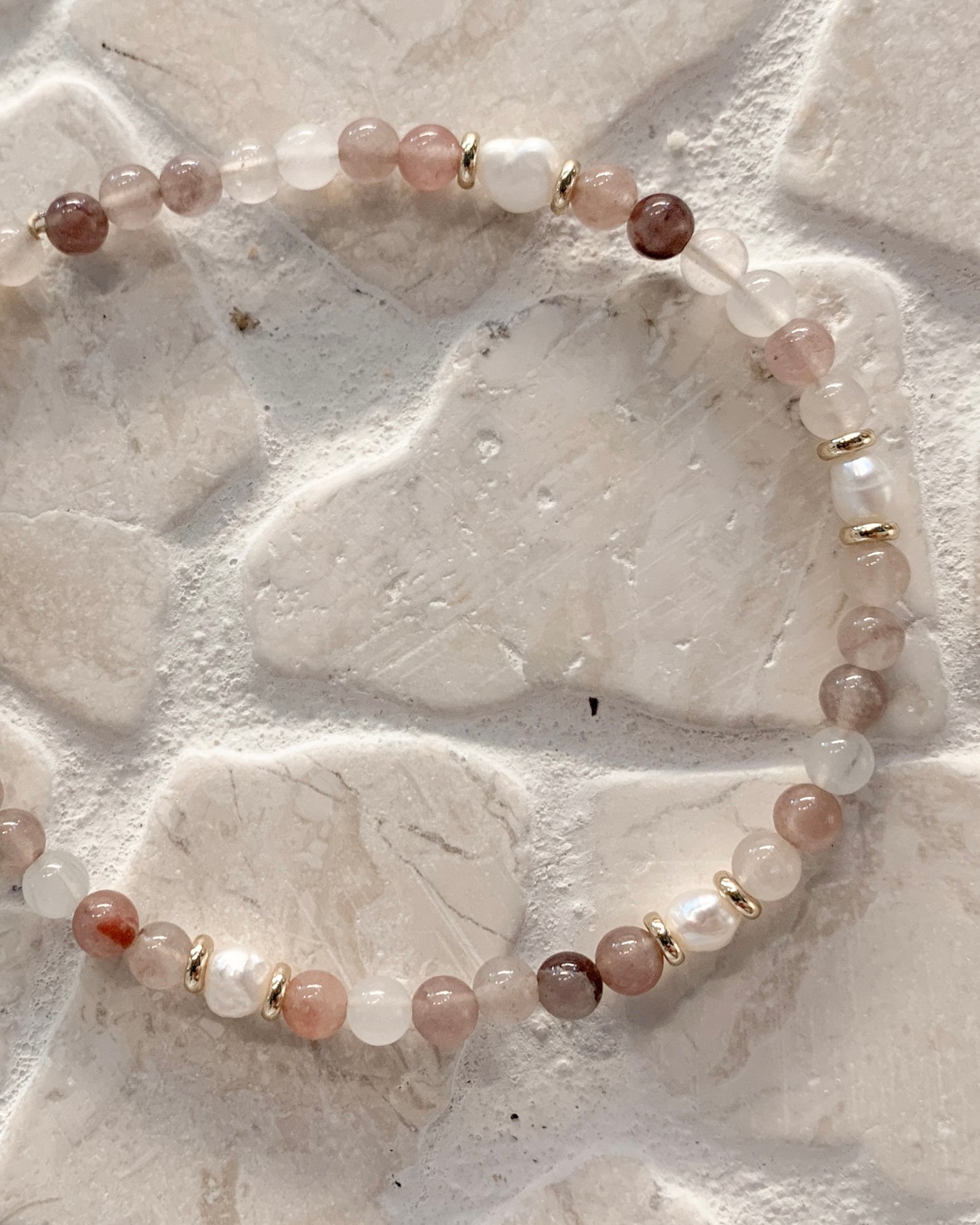 A beaded bracelet made of translucent pink, white, and brown gemstone beads separated by small gold spacer rings, displayed on a textured light beige stone surface.