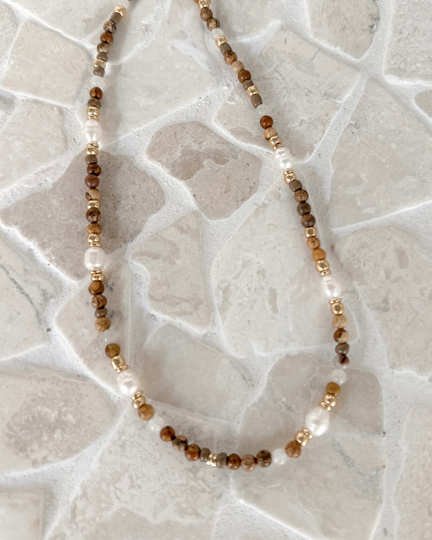 A beaded necklace with brown faceted beads, white pearl-like beads, and small metallic gold spacer beads, arranged on a light stone tile surface.