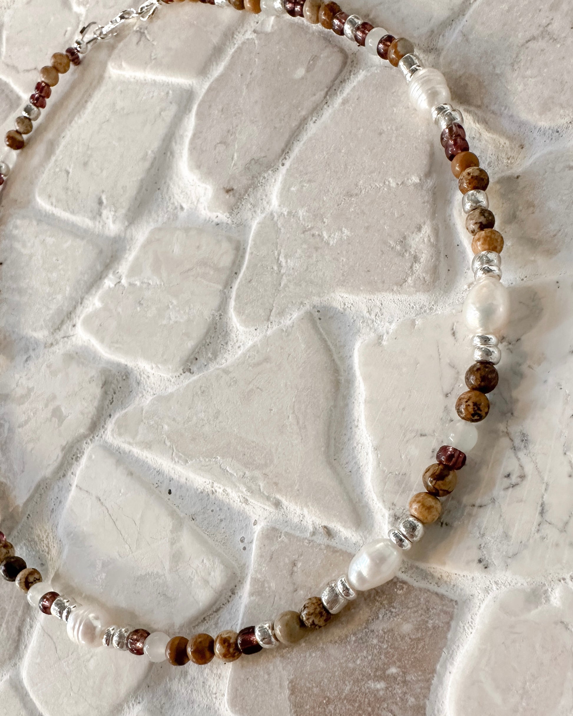 A beaded necklace with picture jasper and  Botswana agate gemstone beads, freshwater pearls and silver-toned spacers, displayed on a textured white stone surface.
