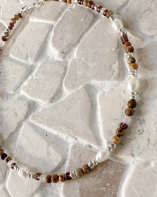 A beaded necklace with picture jasper and  Botswana agate gemstone beads, freshwater pearls and silver-toned spacers, displayed on a textured white stone surface.