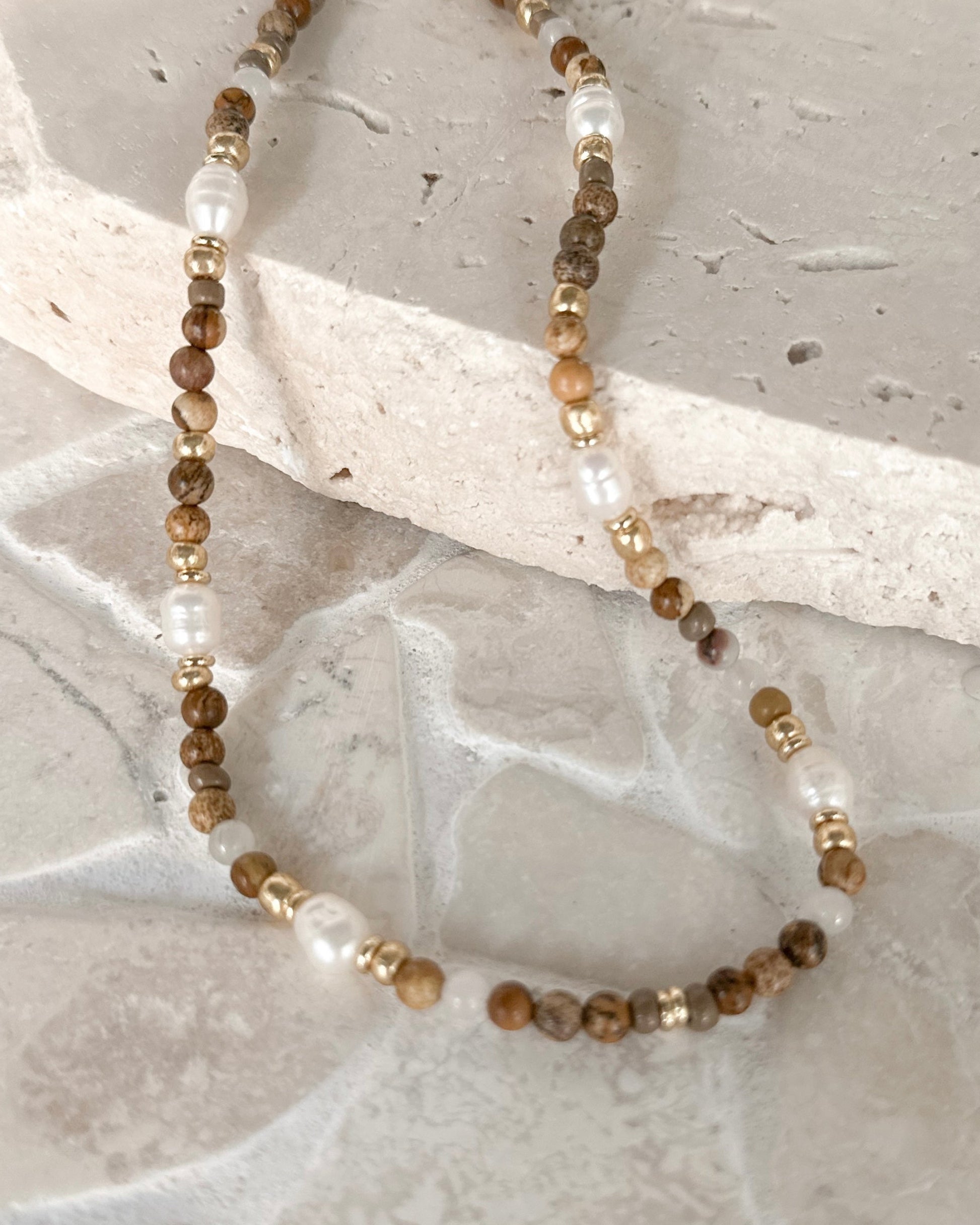 A beaded strand necklace with brown marbled beads, white pearl-like beads, and small gold spacer beads, arranged on a light stone surface.
