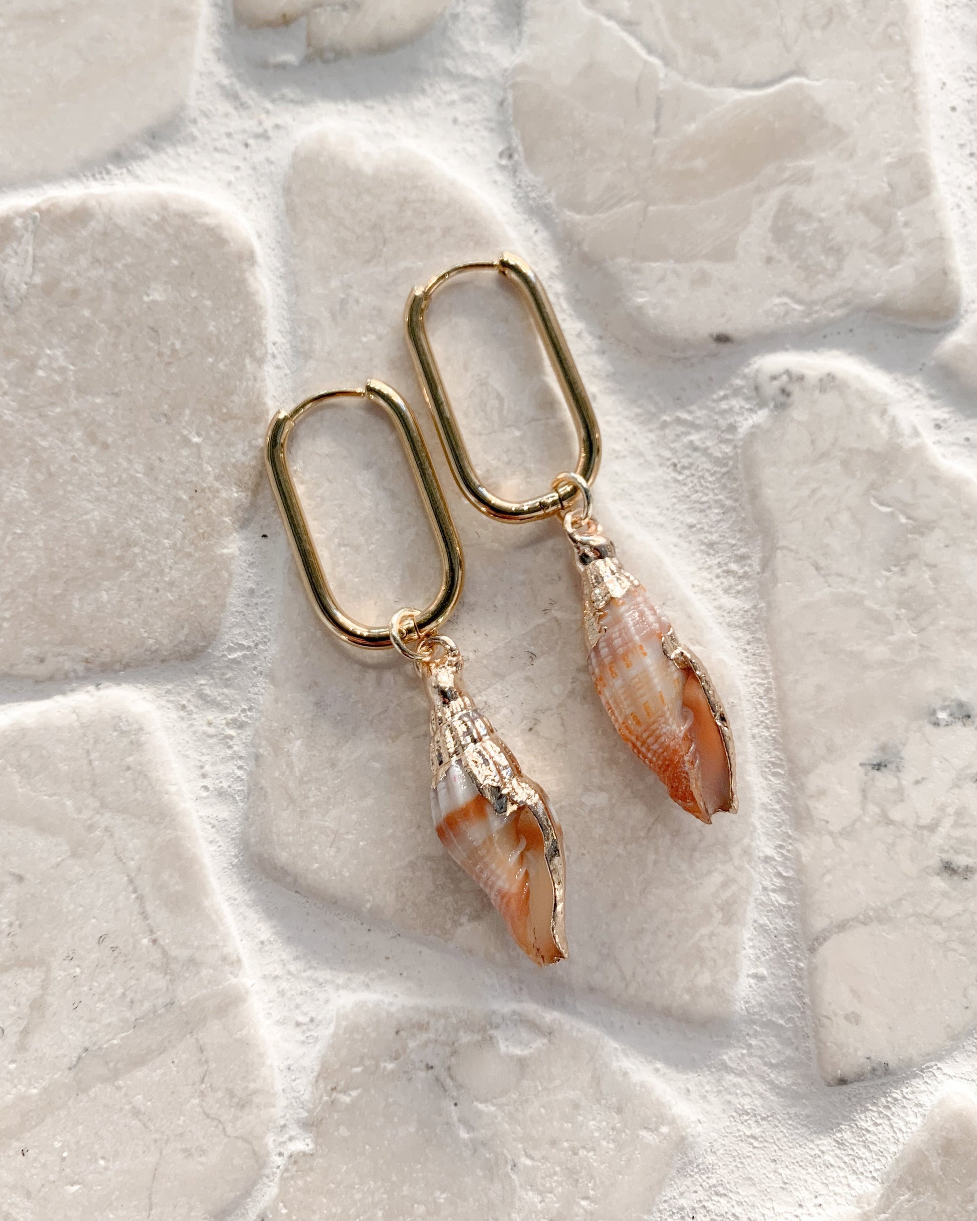 A pair of gold-tone drop earrings featuring conch shell charms, displayed on a textured background of light-colored stone tiles.