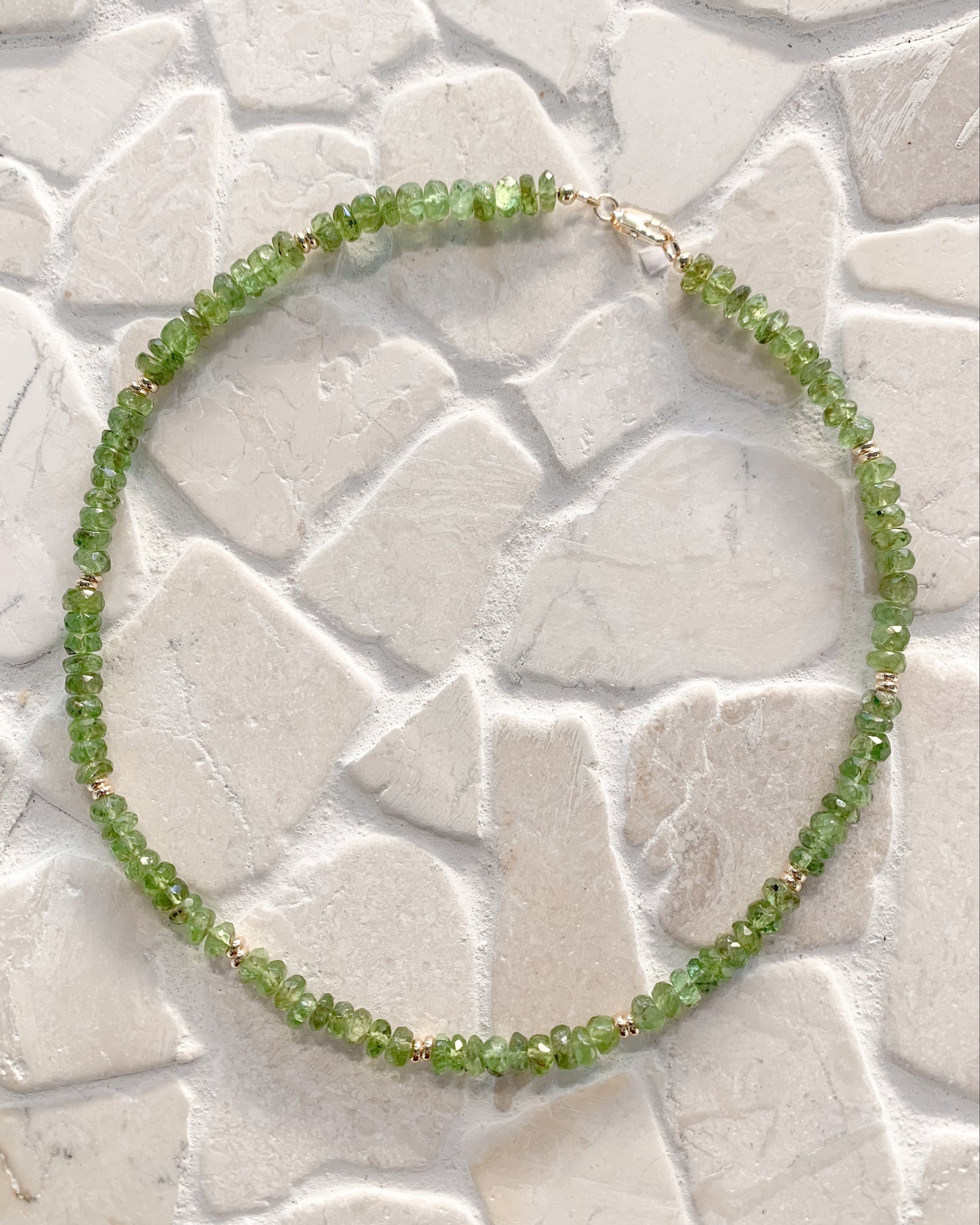 A beaded necklace made of green peridot faceted beads with tiny gold spacer accents, arranged in a circle on a light stone-tile surface.