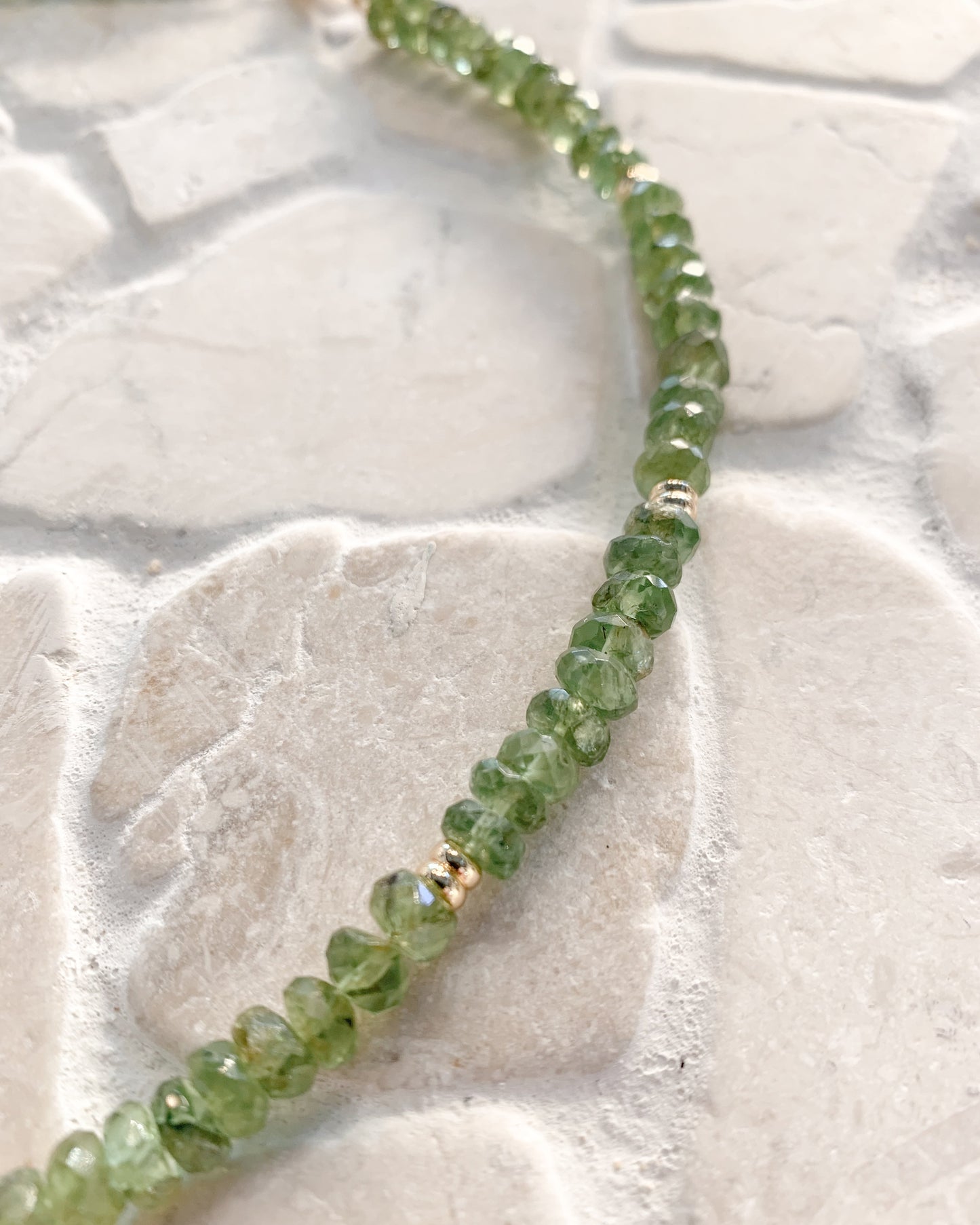A close-up of a beaded peridot strand necklace made of faceted green gemstone beads separated by small metallic spacer beads, laid on a light stone-textured surface.