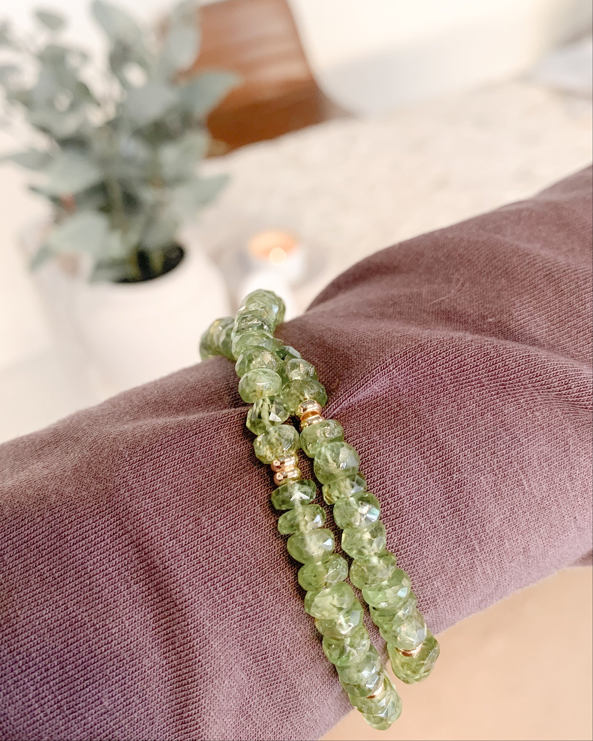 A green faceted peridot gemstone beaded necklace with small gold spacer beads, shown wrapped on a person's wrist wrapped in a mauve fabric sleeve.