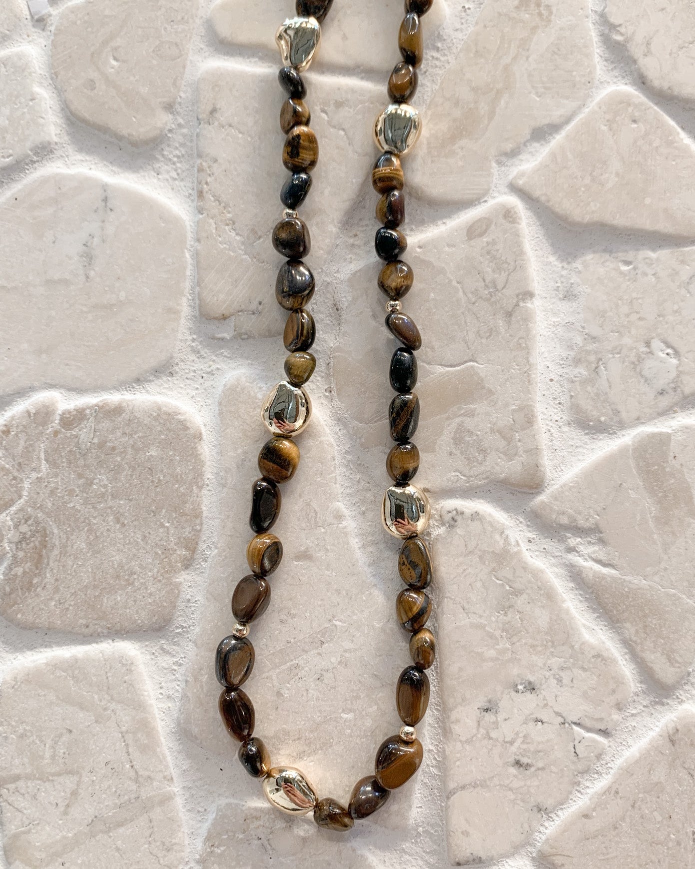 A beaded necklace made of polished tiger's eye beads with a few clear faceted accent beads, displayed on a light beige stone tile background.