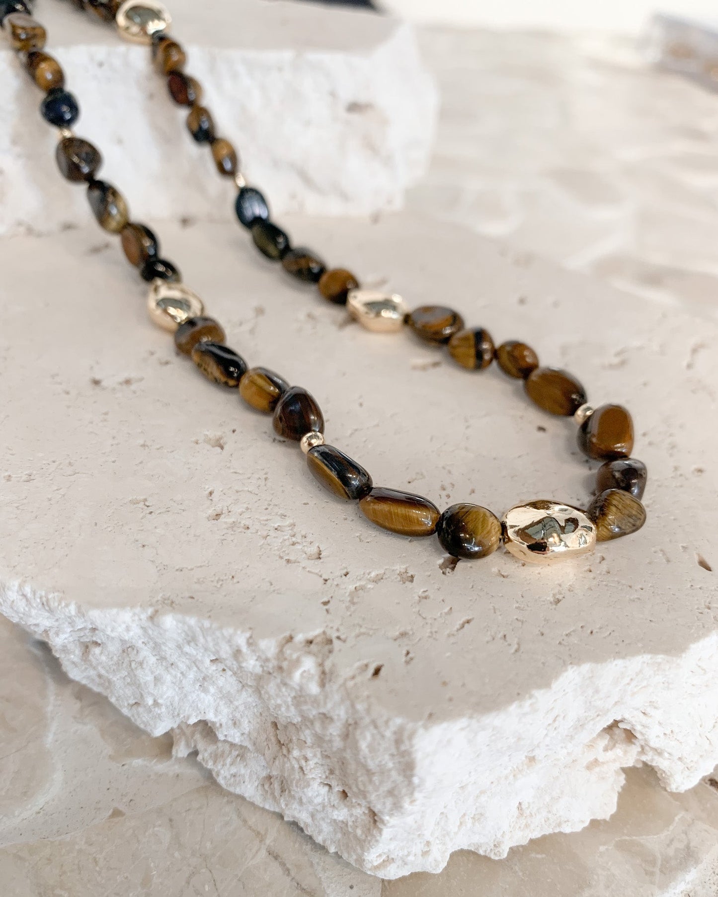A beaded necklace with polished brown and black tiger's eye stones and a few metallic gold spacer beads, displayed on a textured white stone slab.