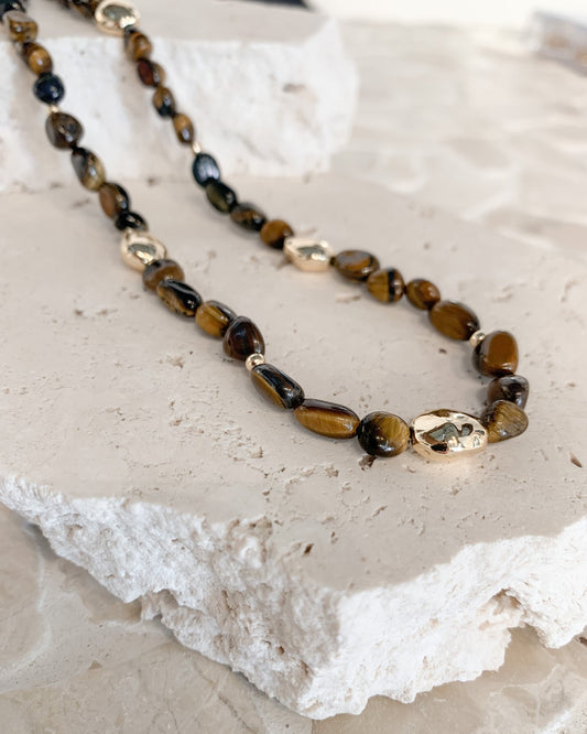 A beaded necklace with polished brown and black tiger's eye stones and a few metallic gold spacer beads, displayed on a textured white stone slab.