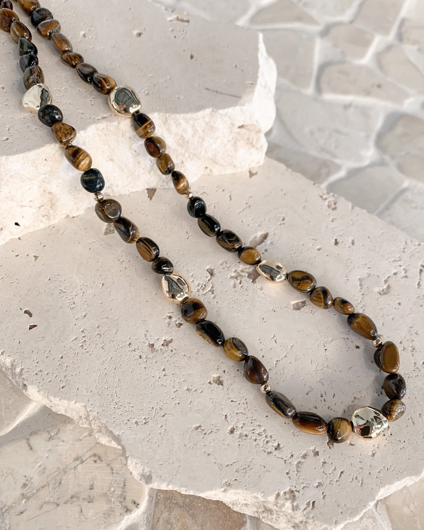 A beaded necklace made of polished tiger's eye and clear faceted beads, displayed on a textured white stone slab over a light stone tile surface.