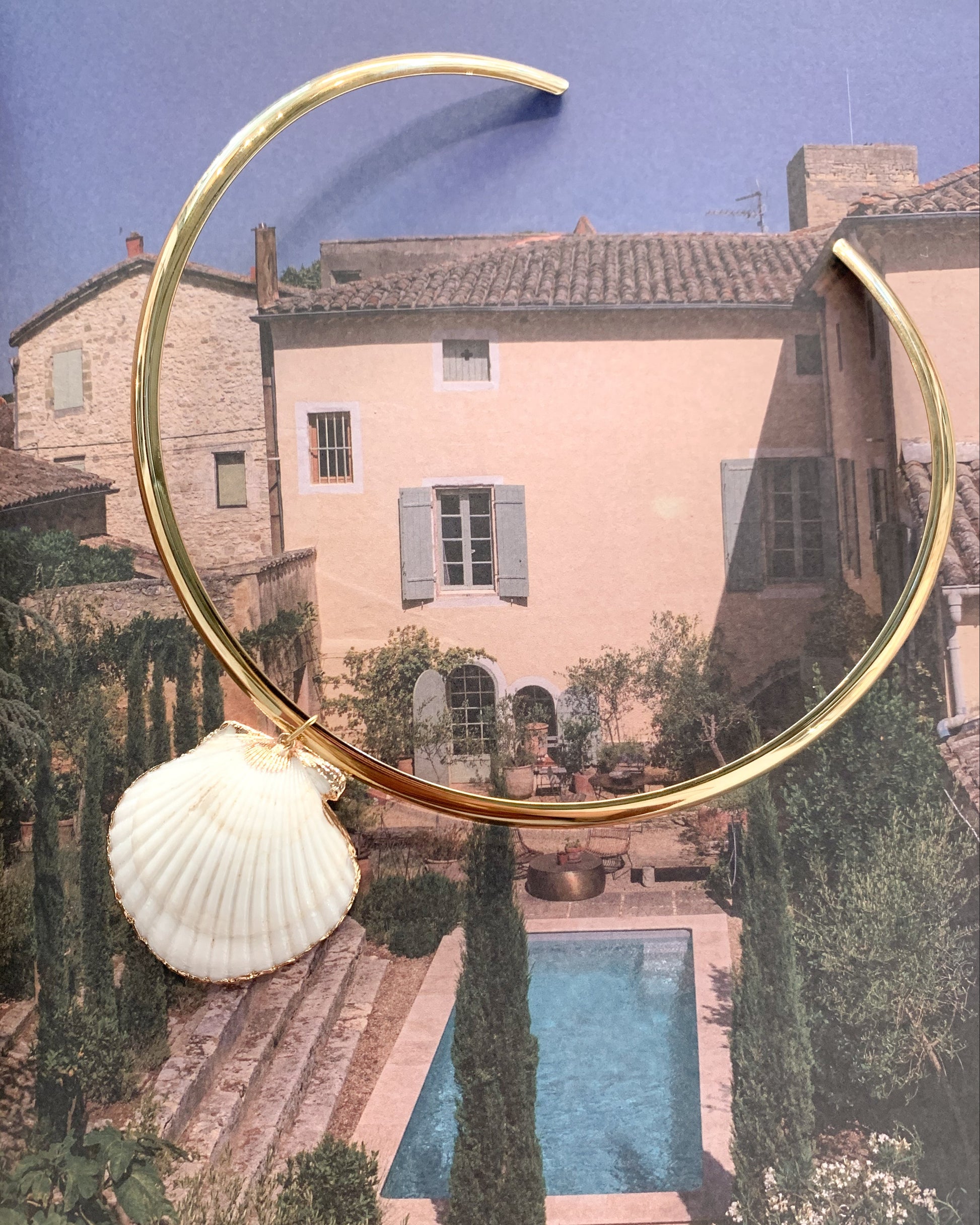 A gold crescent-shaped collar necklace with a white scallop shell charm, displayed over a scenic photograph of a Mediterranean villa and pool.