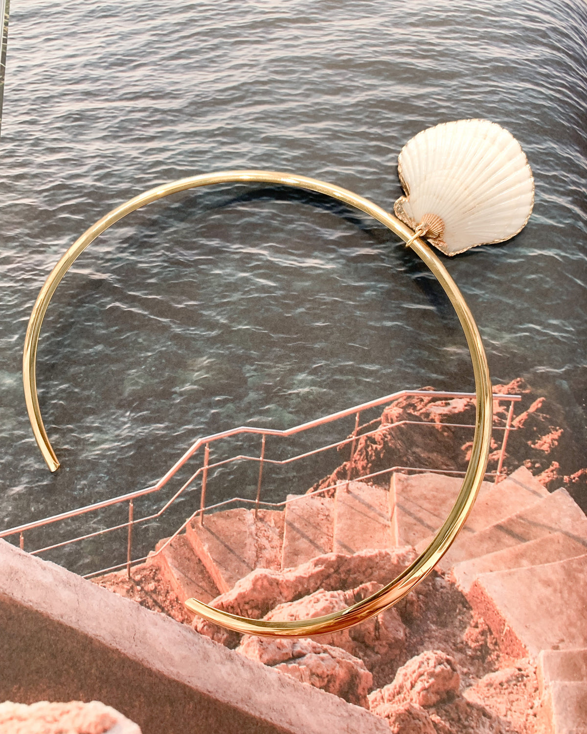 A gold collar necklace with a white seashell charm, displayed against a scenic background of ocean water, terracotta stairs, and a stone wall.