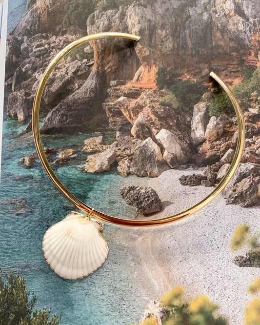 A gold-tone collar necklace with a white scallop shell pendant, displayed against a scenic beach and rocky coastline background.
