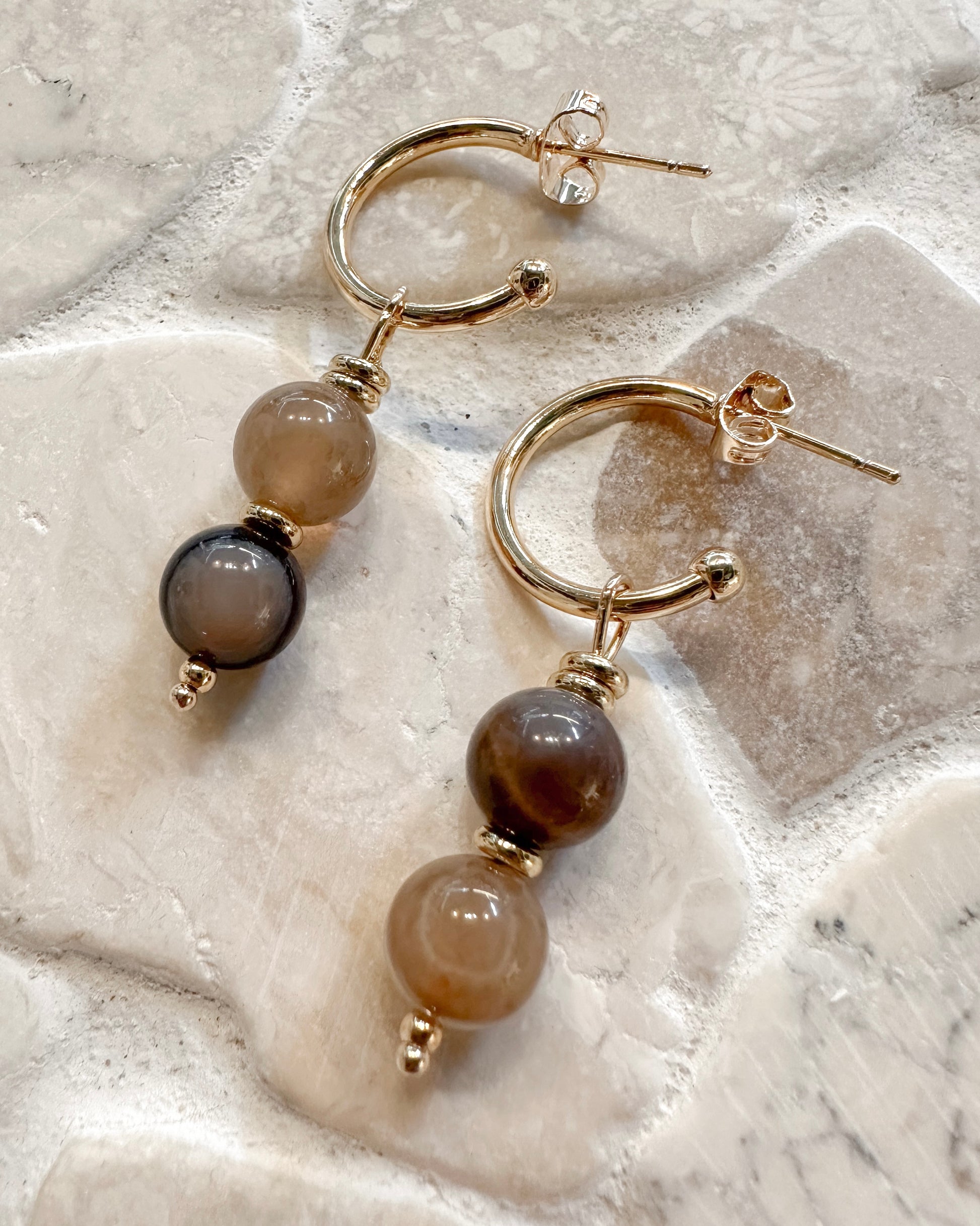 Gold hoop drop earrings with smooth round beads in taupe, brown, and black, displayed on a textured light stone surface.