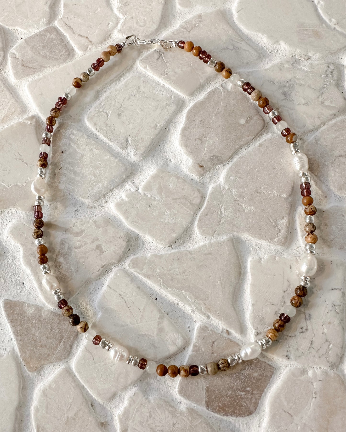 A beaded necklace with picture jasper and  Botswana agate gemstone beads, freshwater pearls and silver-toned spacers, displayed on a textured white stone surface.