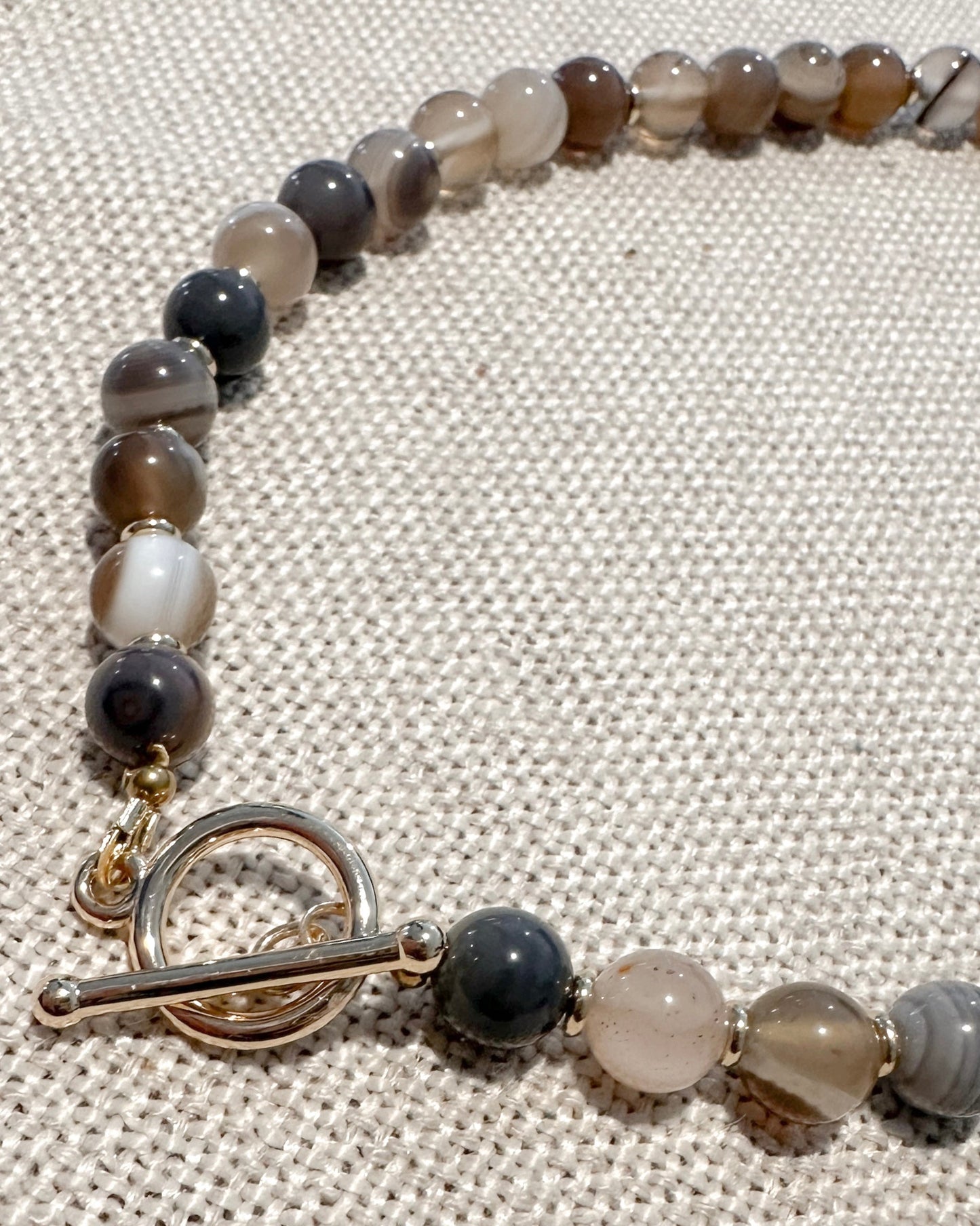 A beaded necklace with polished agate-like round beads in earthy gray, brown, white, and black tones, featuring a gold-tone toggle clasp, displayed on a textured beige fabric background.