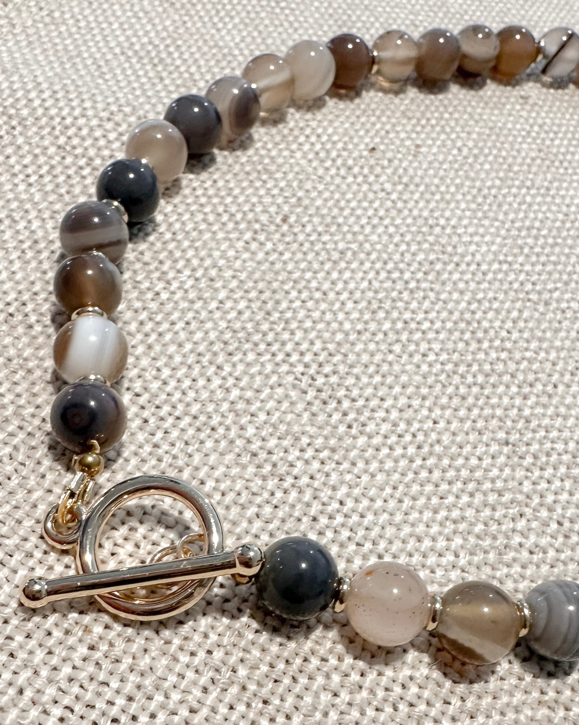 A beaded necklace with polished agate-like round beads in earthy gray, brown, white, and black tones, featuring a gold-tone toggle clasp, displayed on a textured beige fabric background.