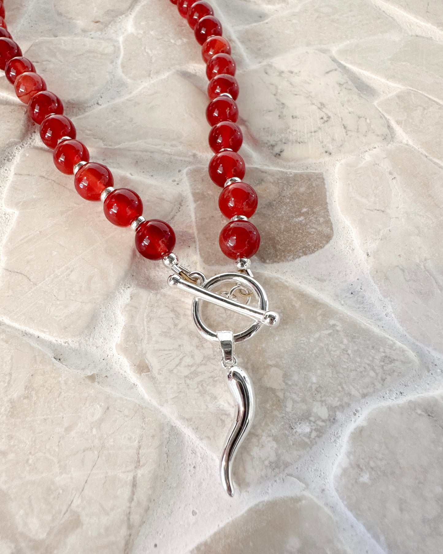 A beaded necklace made of polished red carnelian beads with silver spacer rings and a silver toggle clasp, displayed on a light beige stone tile surface.