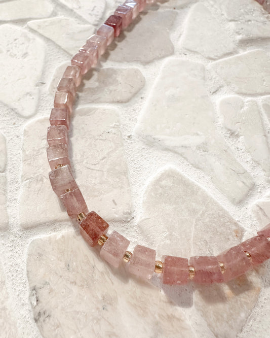 A strand of chunky, faceted pink gemstone beads separated by small gold spacer rings, laid out on a light beige stone tile background.