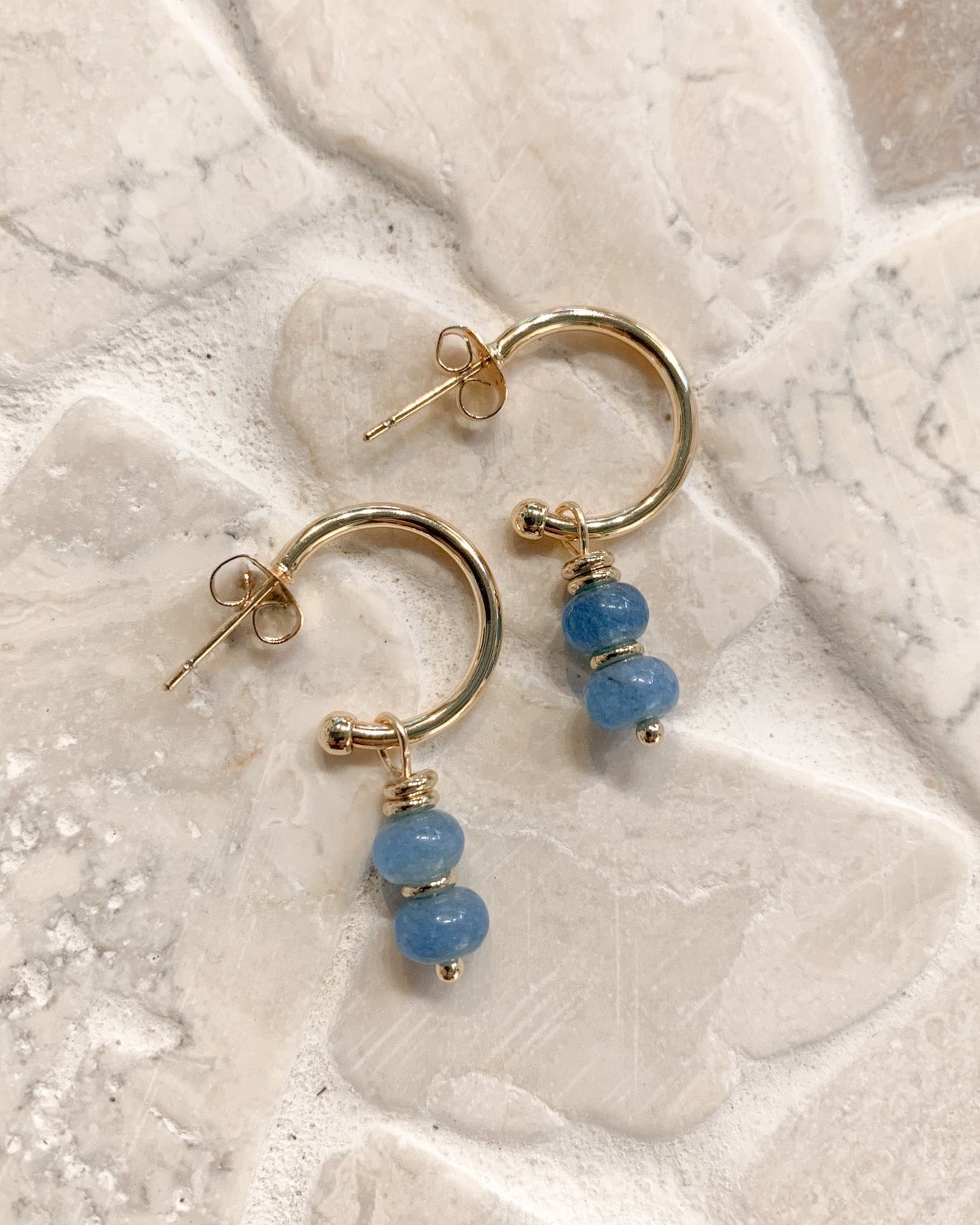 Gold hoop earrings with two light blue beads dangling from each hoop, displayed on a light beige stone tile background.