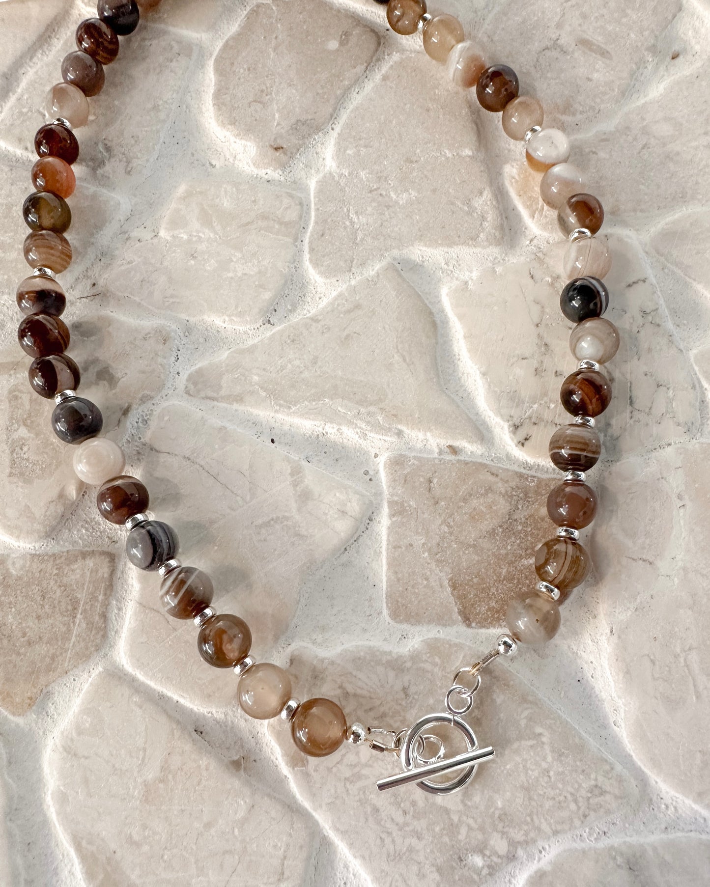 A beaded necklace made of polished round gemstone beads in earthy brown, smoky gray, cream, and black, with small silver spacer beads and a silver toggle clasp, displayed on a light beige stone tile background.