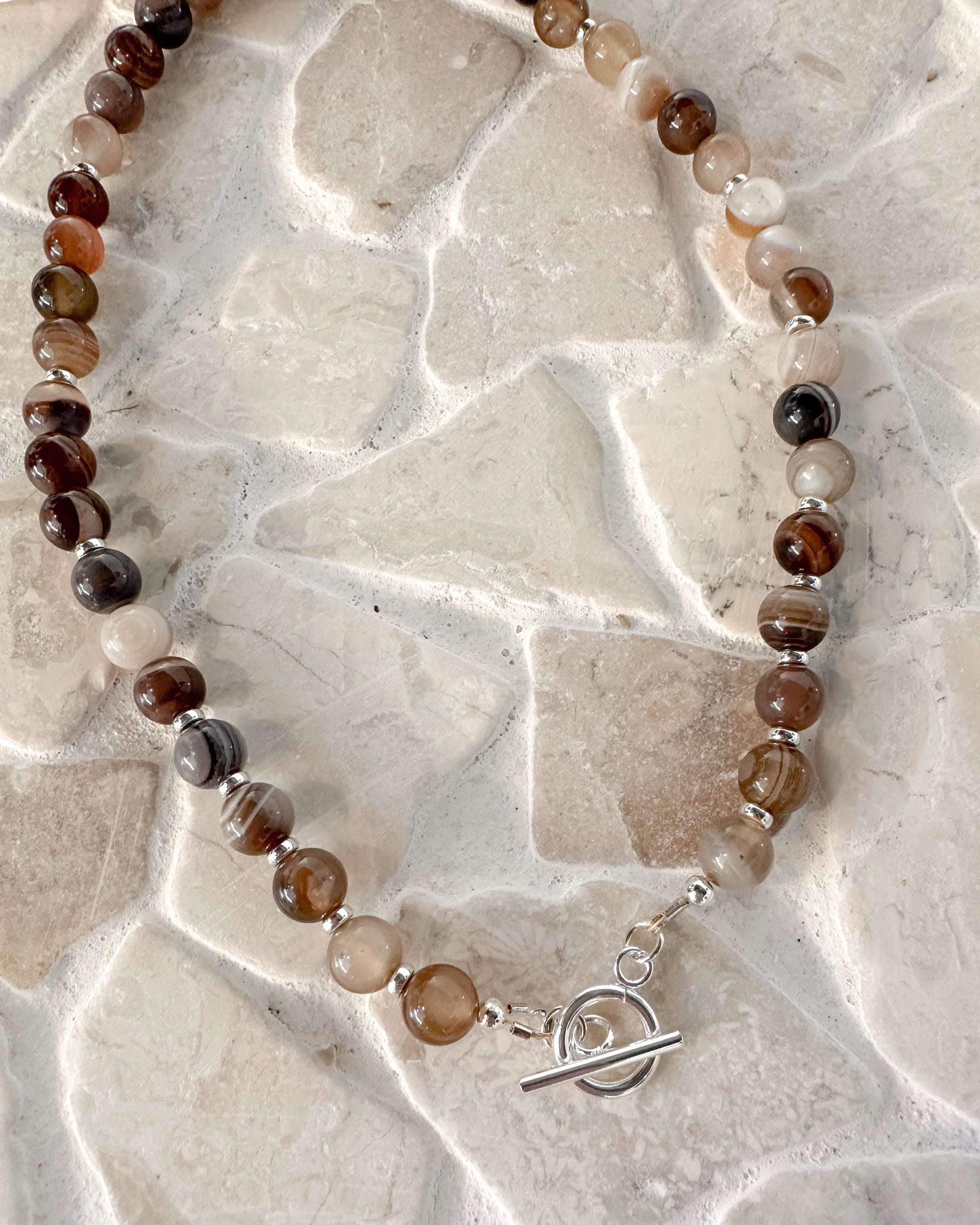 A beaded necklace made of polished round gemstone beads in earthy brown, smoky gray, cream, and black, with small silver spacer beads and a silver toggle clasp, displayed on a light beige stone tile background.